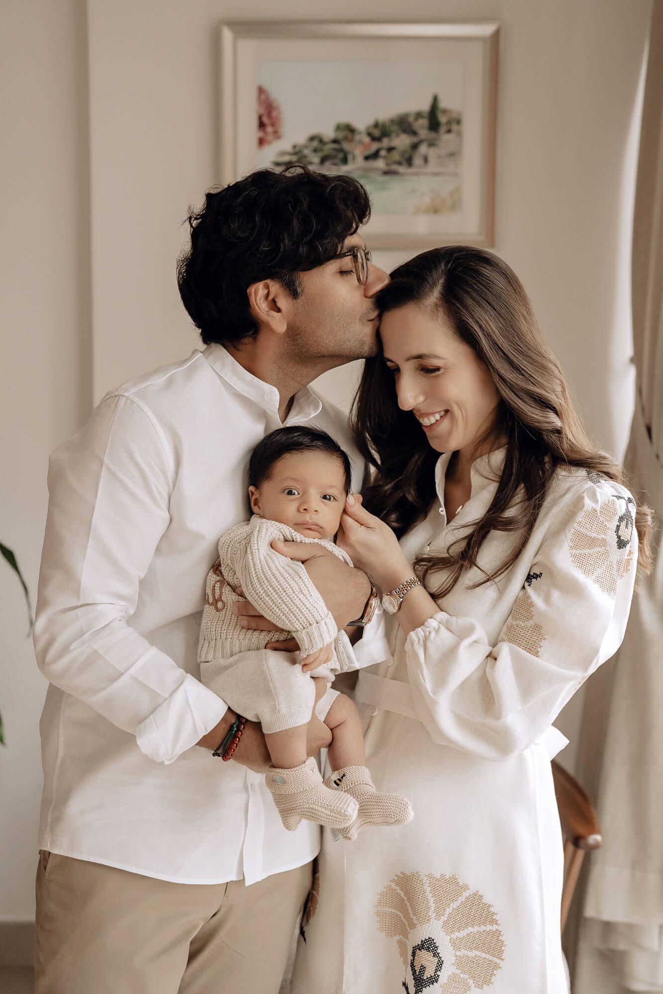 Intimate family portrait showing tender moment between parents and baby in matching white outfits.