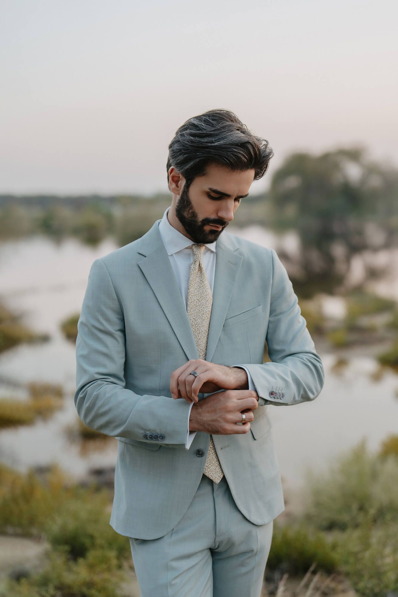 Stylish light blue suit with subtle details and cream tie pictured against a serene outdoor backdrop at dusk.