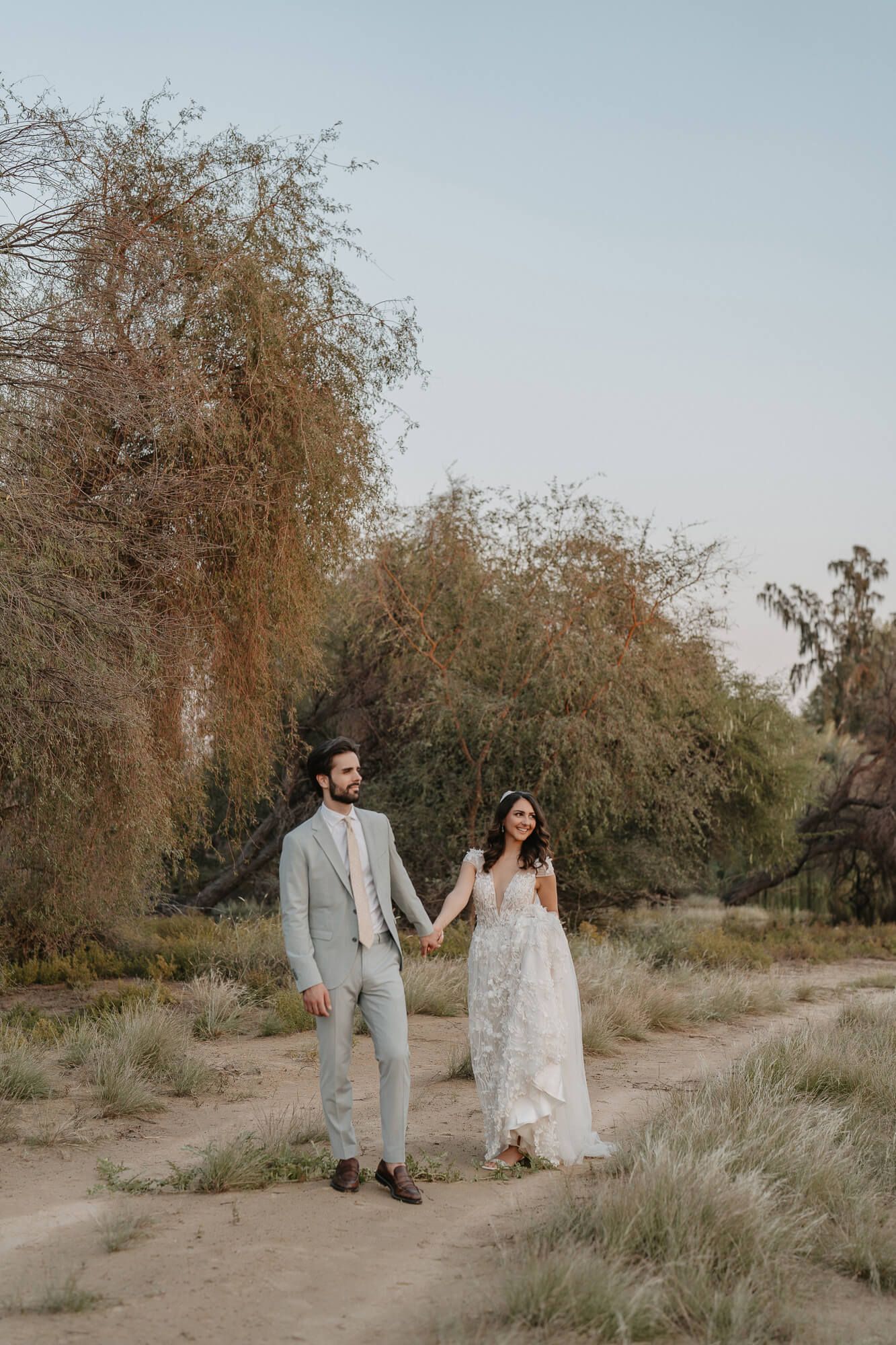 A couple holds hands on a desert path with willow trees and soft lighting creating a romantic outdoor scene.