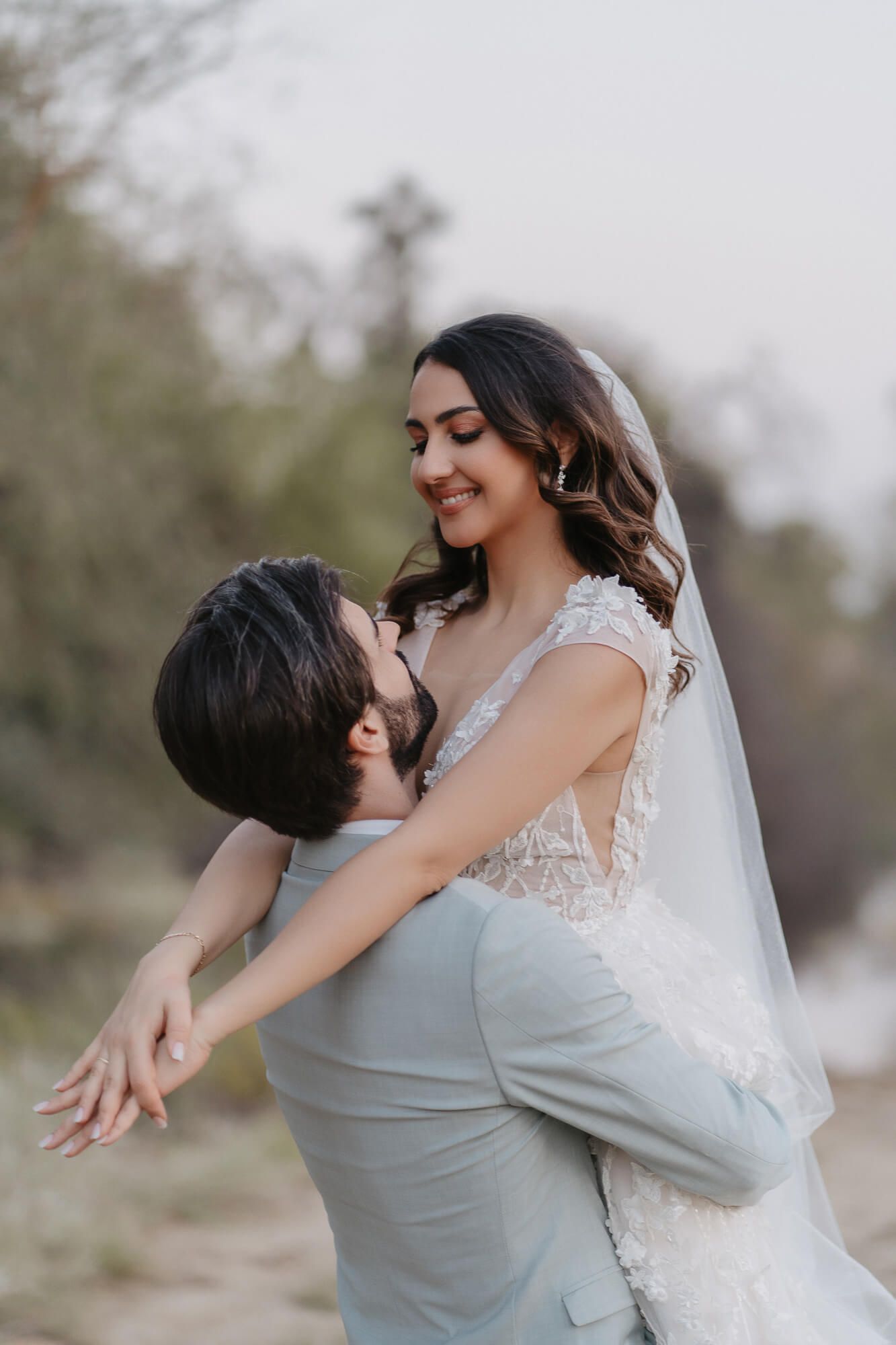 A romantic wedding couple shares an intimate moment outdoors with the bride wearing a flowing white lace gown and veil.