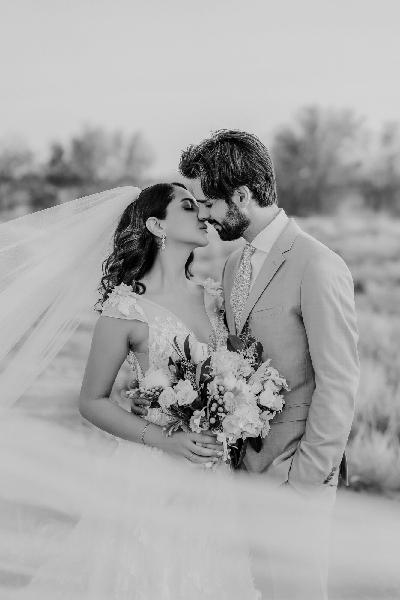 Romantic black and white wedding photo showing newlyweds sharing an intimate moment with bridal bouquet in an outdoor setting.