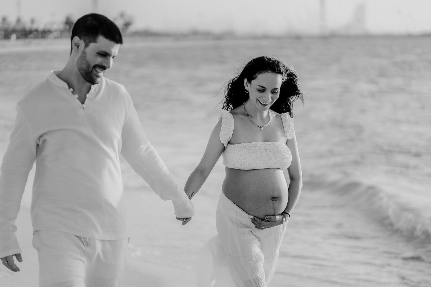 Black and white photo of an expectant couple walking hand in hand on a beach wearing light colored clothing.