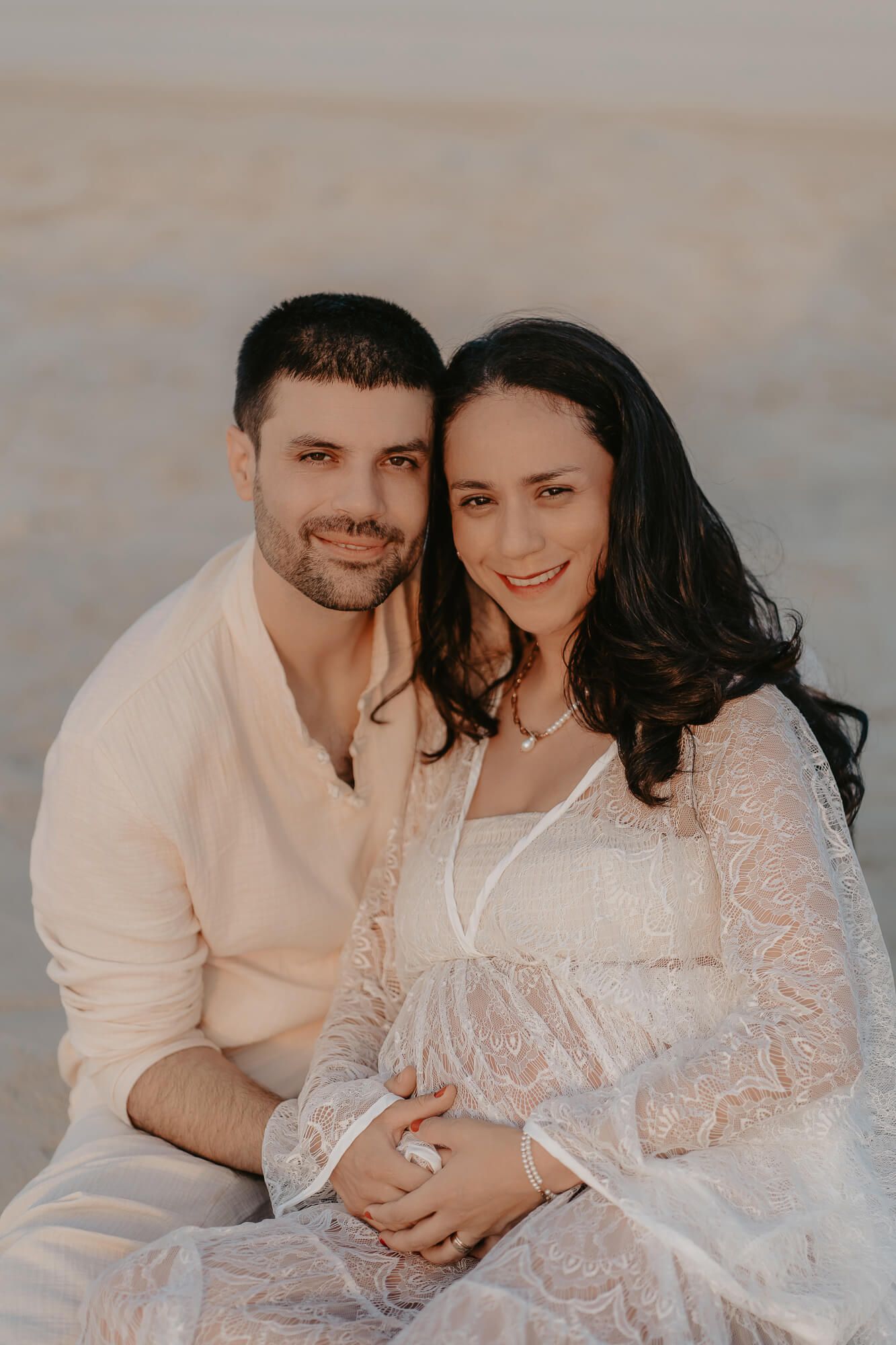 A romantic couple sits on a beach in coordinated white outfits during sunset for their engagement photos.