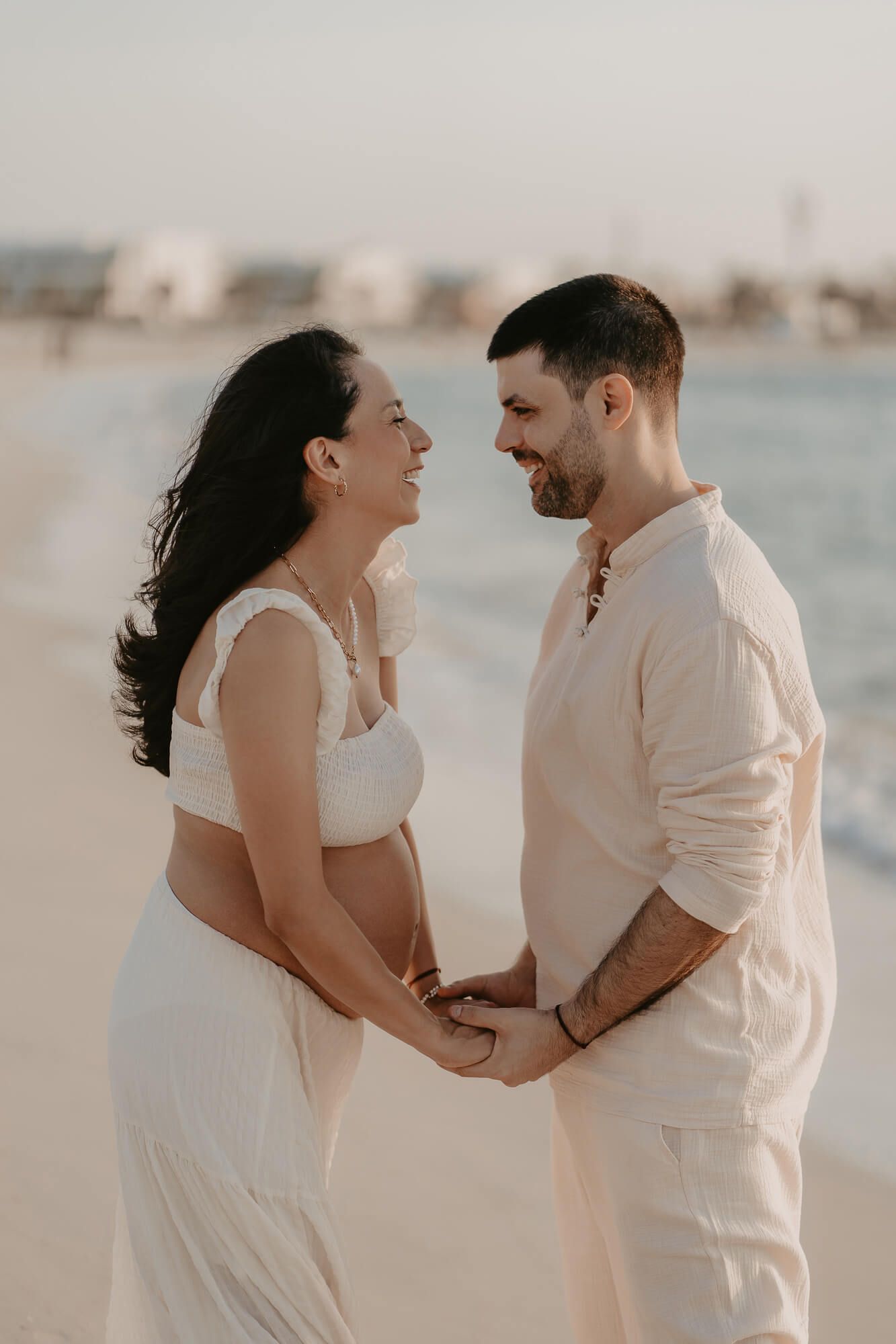 A couple in white outfits share a romantic moment while holding hands on a beach at sunset.