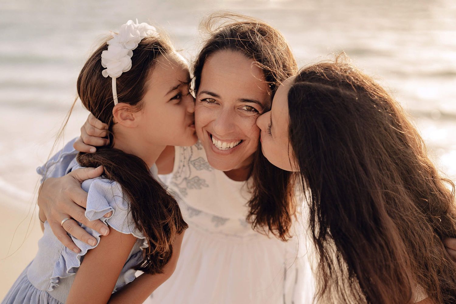 Three people sharing an intimate moment of laughter and kisses on a beach at sunset.