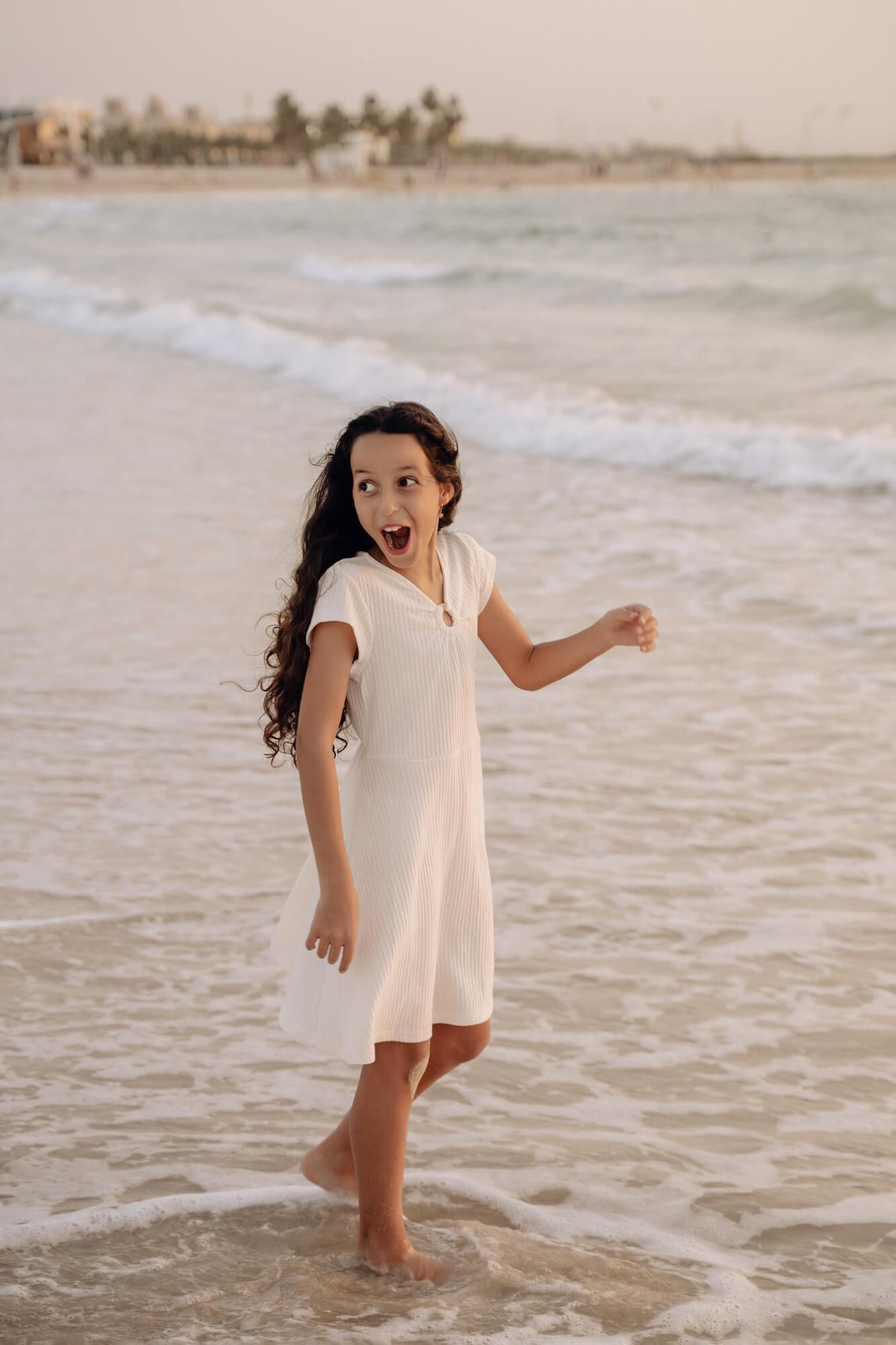 A person in a white summer dress plays joyfully in the ocean waves at sunset.