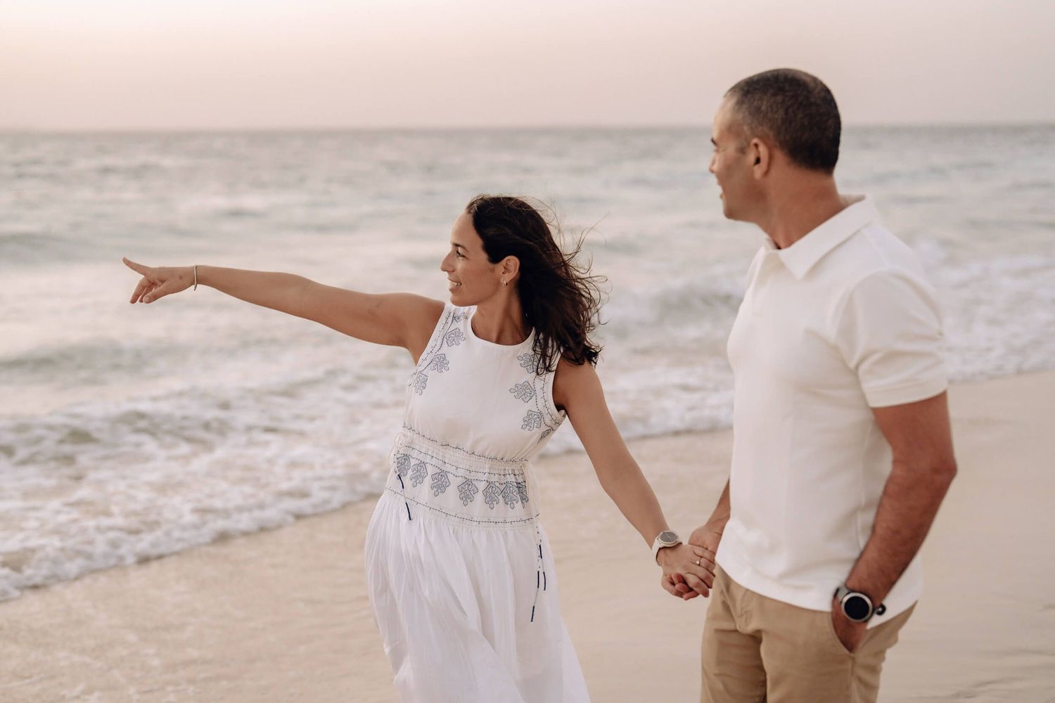 Couple walking hand in hand on beach while one points toward the ocean.