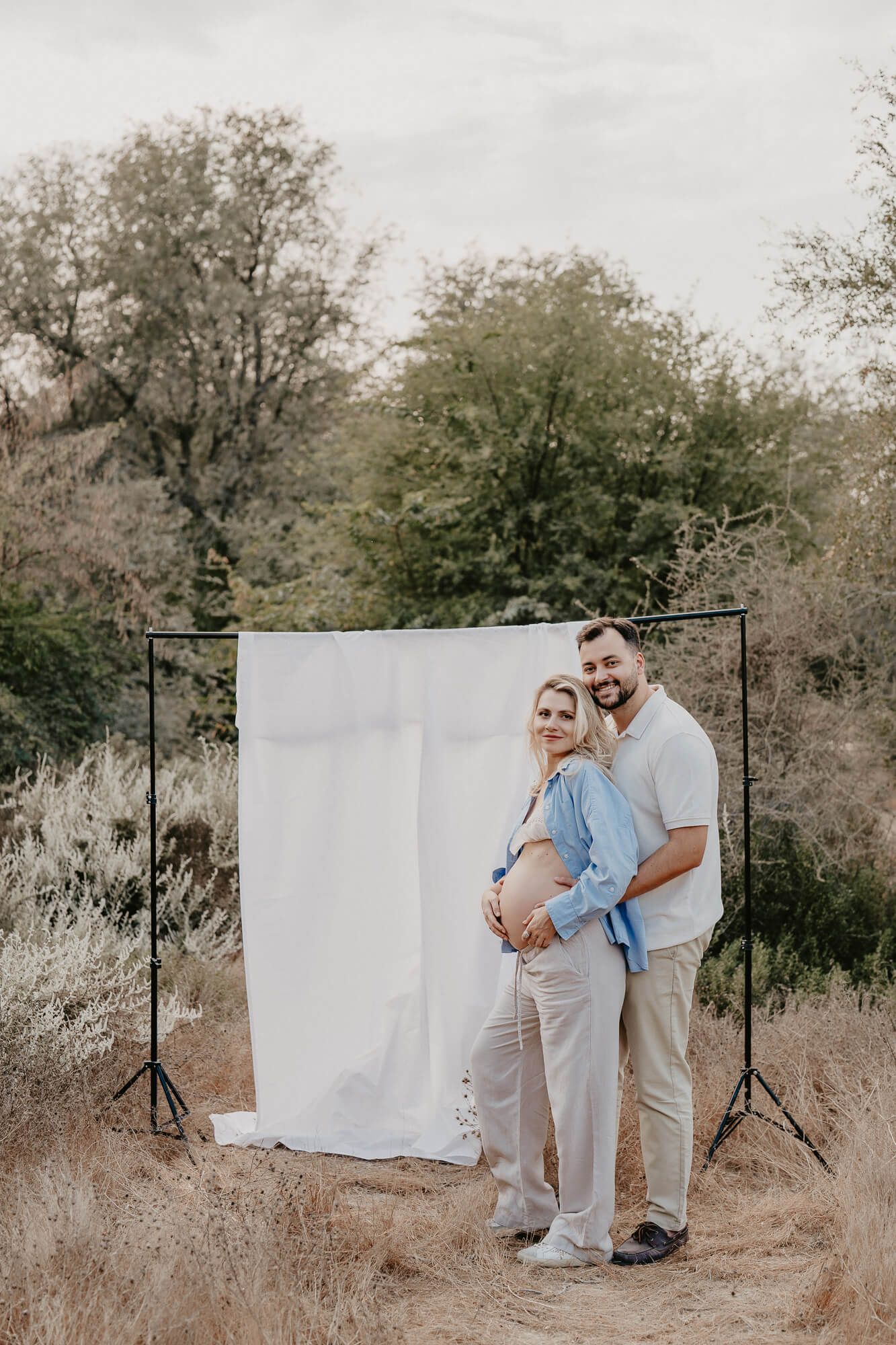 Expecting couple posing together against white backdrop in outdoor setting with trees.