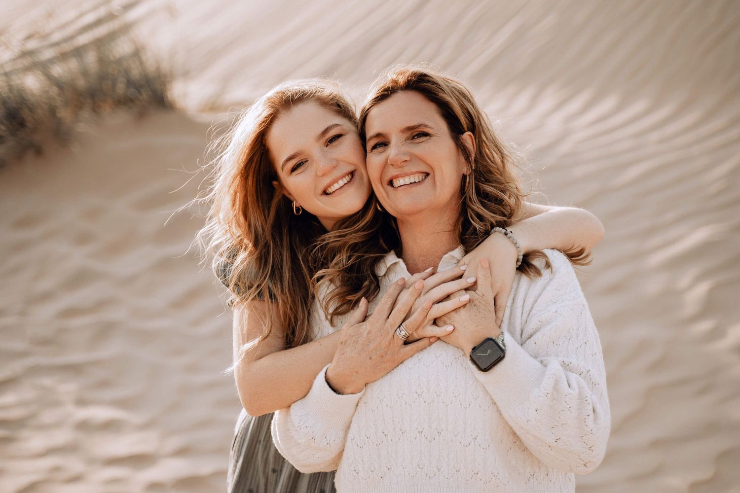 Two people share heartfelt embrace outdoors with sand dunes in background.