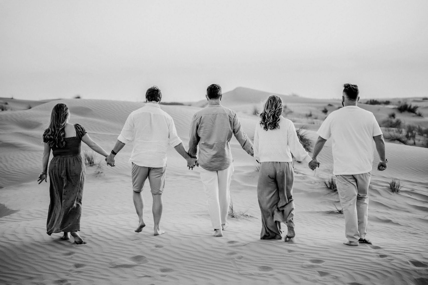 Five people hold hands while walking along desert sand dunes in black and white.