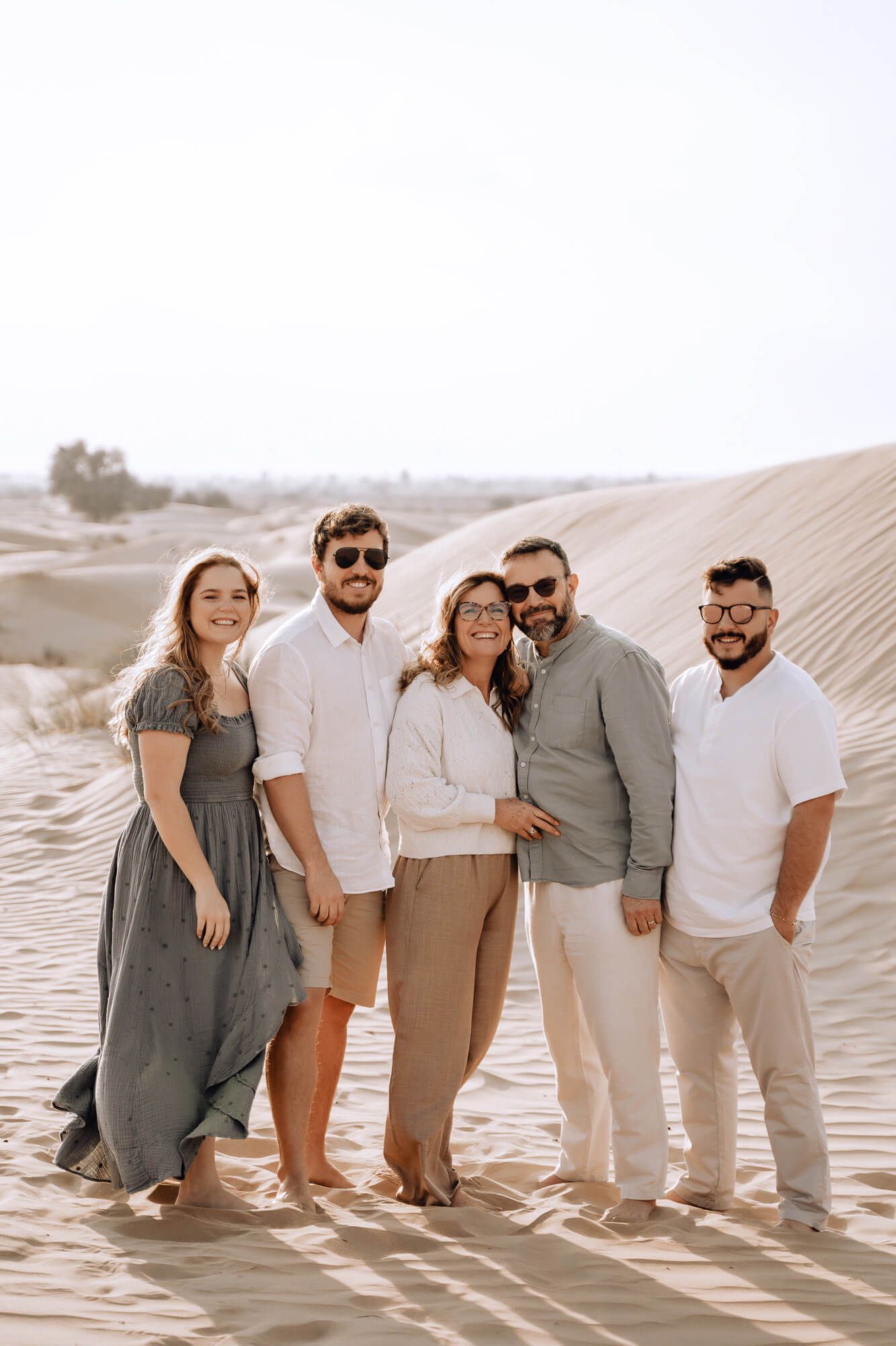 Five people wearing coordinated cream and gray outfits stand together smiling on desert sand dunes.