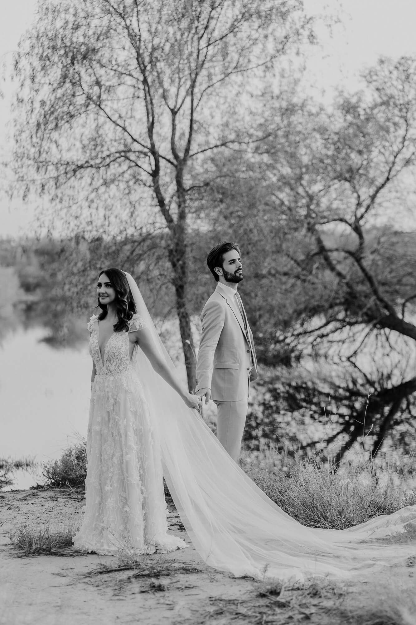 A wedding couple holds hands in a dreamy black and white photo by a lake with trees in the background.