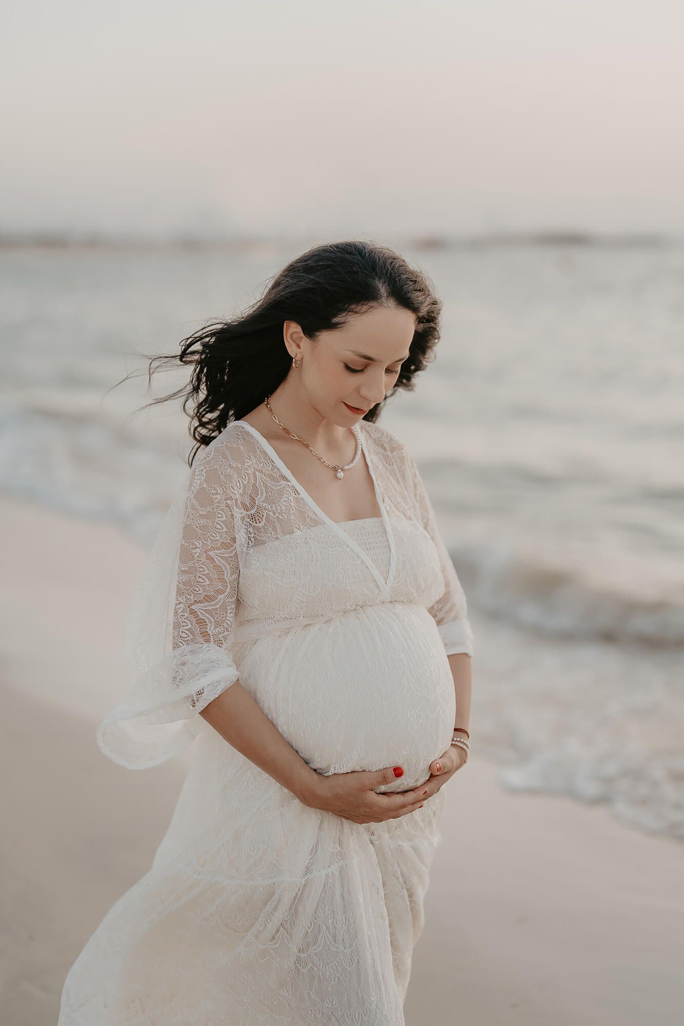 Ethereal maternity portrait on beach with white lace dress and soft lighting during sunset.