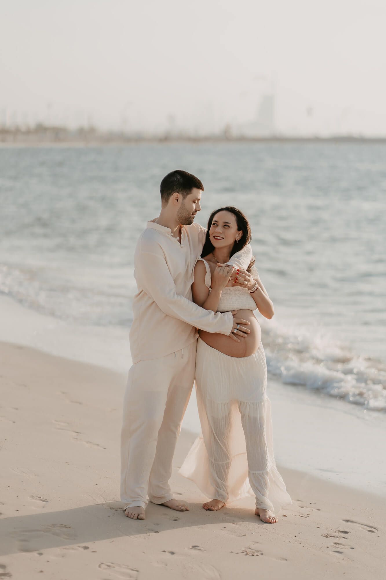 A couple wearing white outfits sits together on a sandy beach with ocean waves in the background.