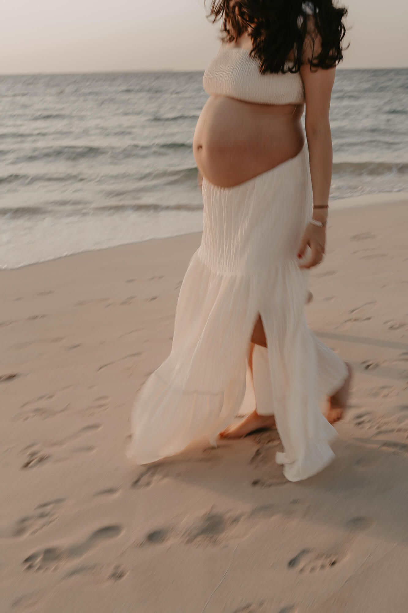 A person in a flowing white dress walks barefoot along a sandy beach at sunset.