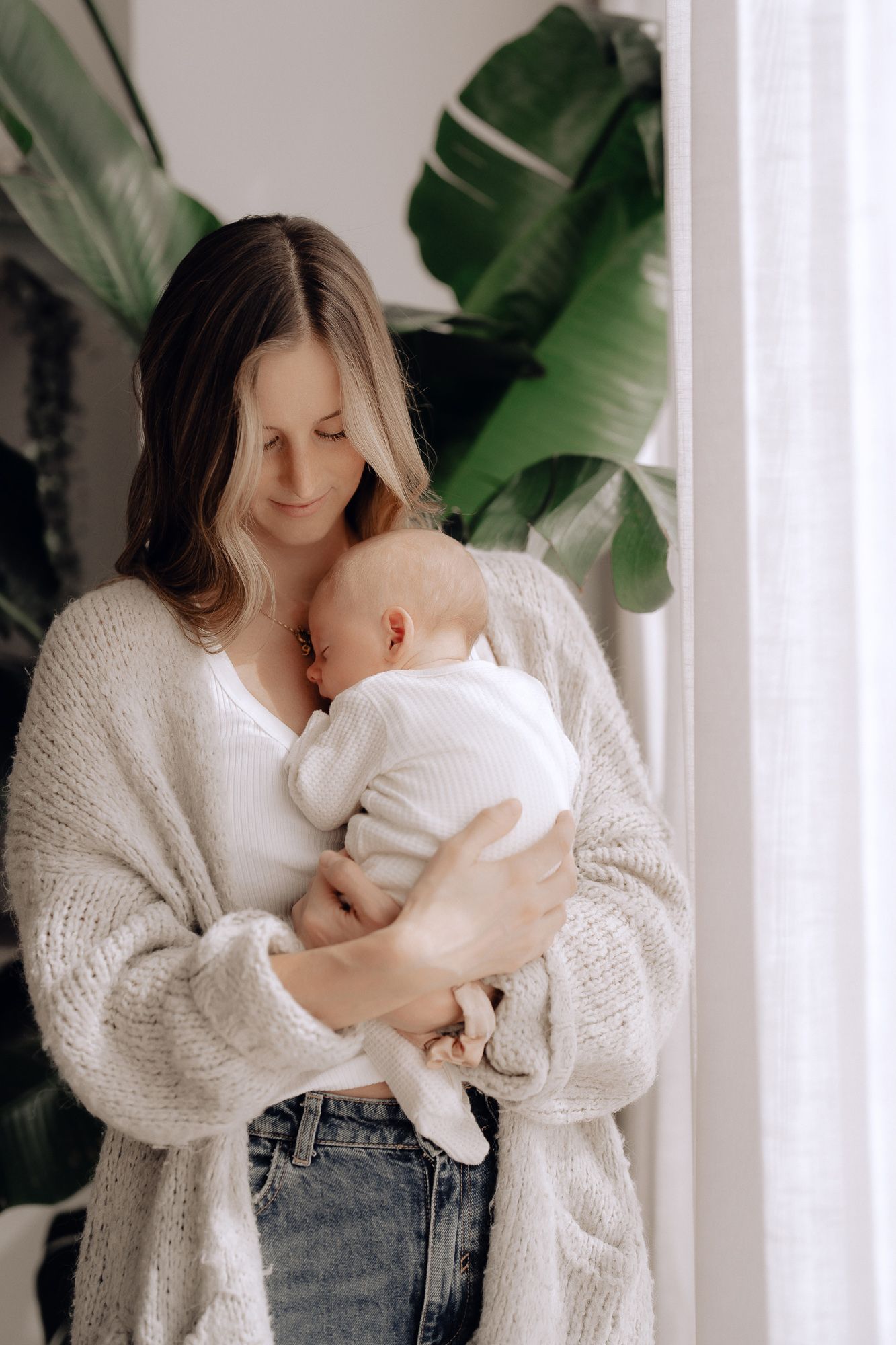 Tender moment between parent and baby in white knit cardigan by large indoor plant leaves against bright window.