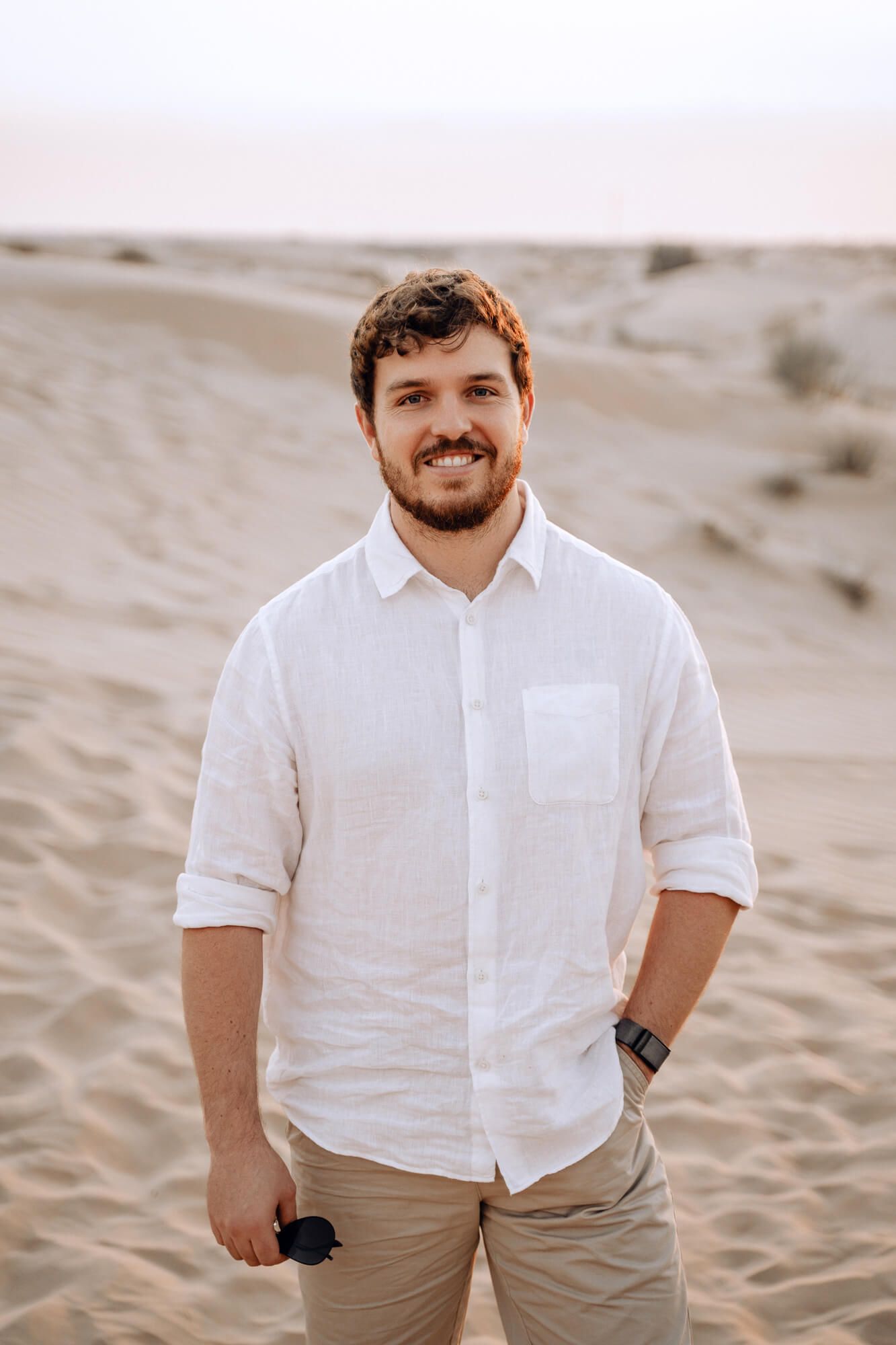 Person in white button-down shirt smiles warmly while standing in desert sand dunes.