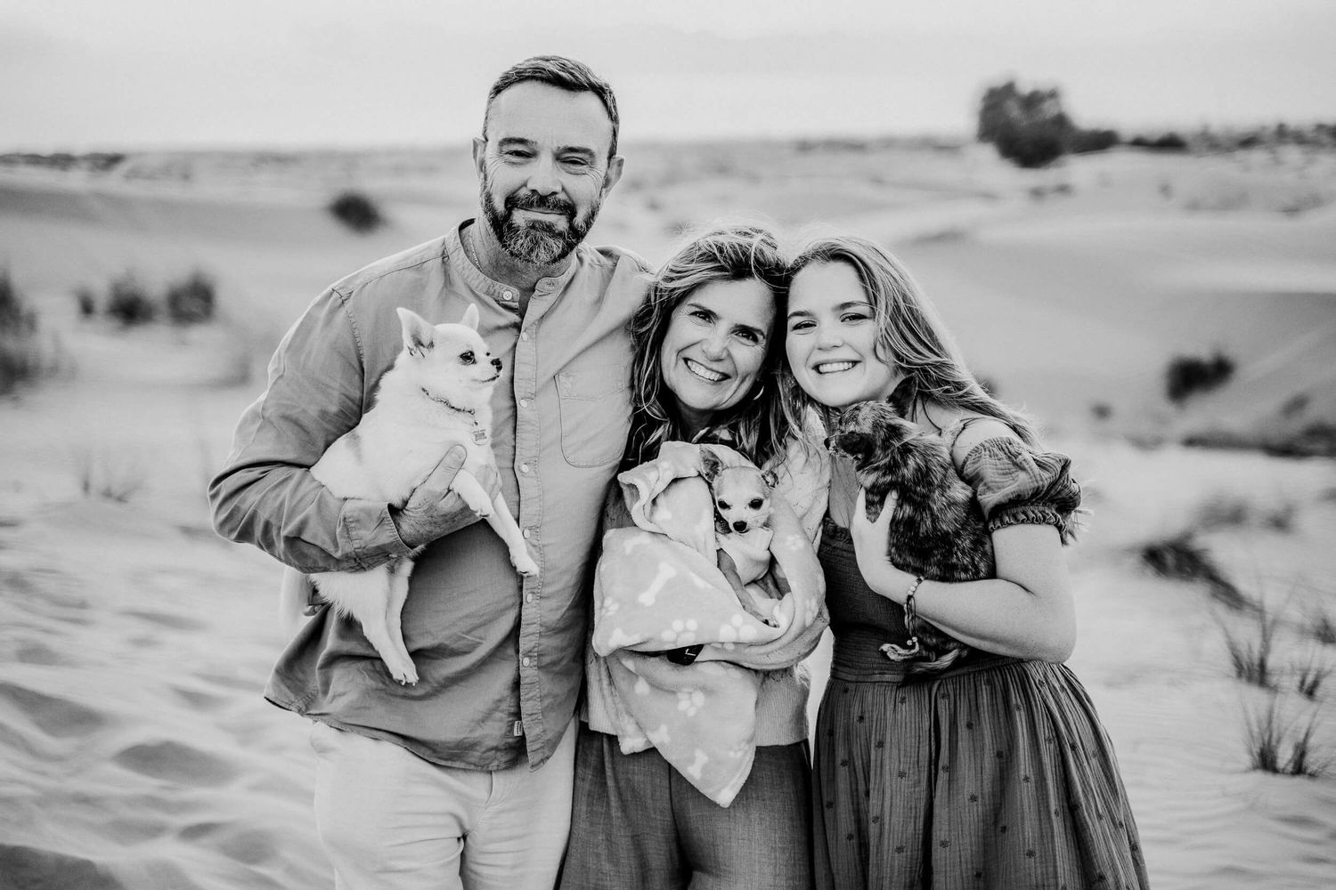 Couple embracing their chihuahua while sitting in desert sand dunes.