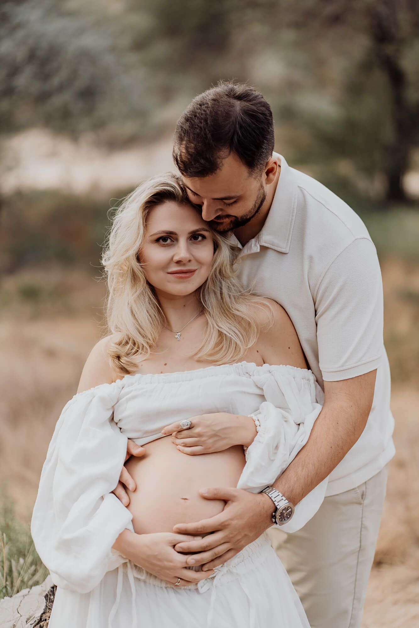 Romantic maternity photo series with couple in coordinated white and beige outfits outdoors.