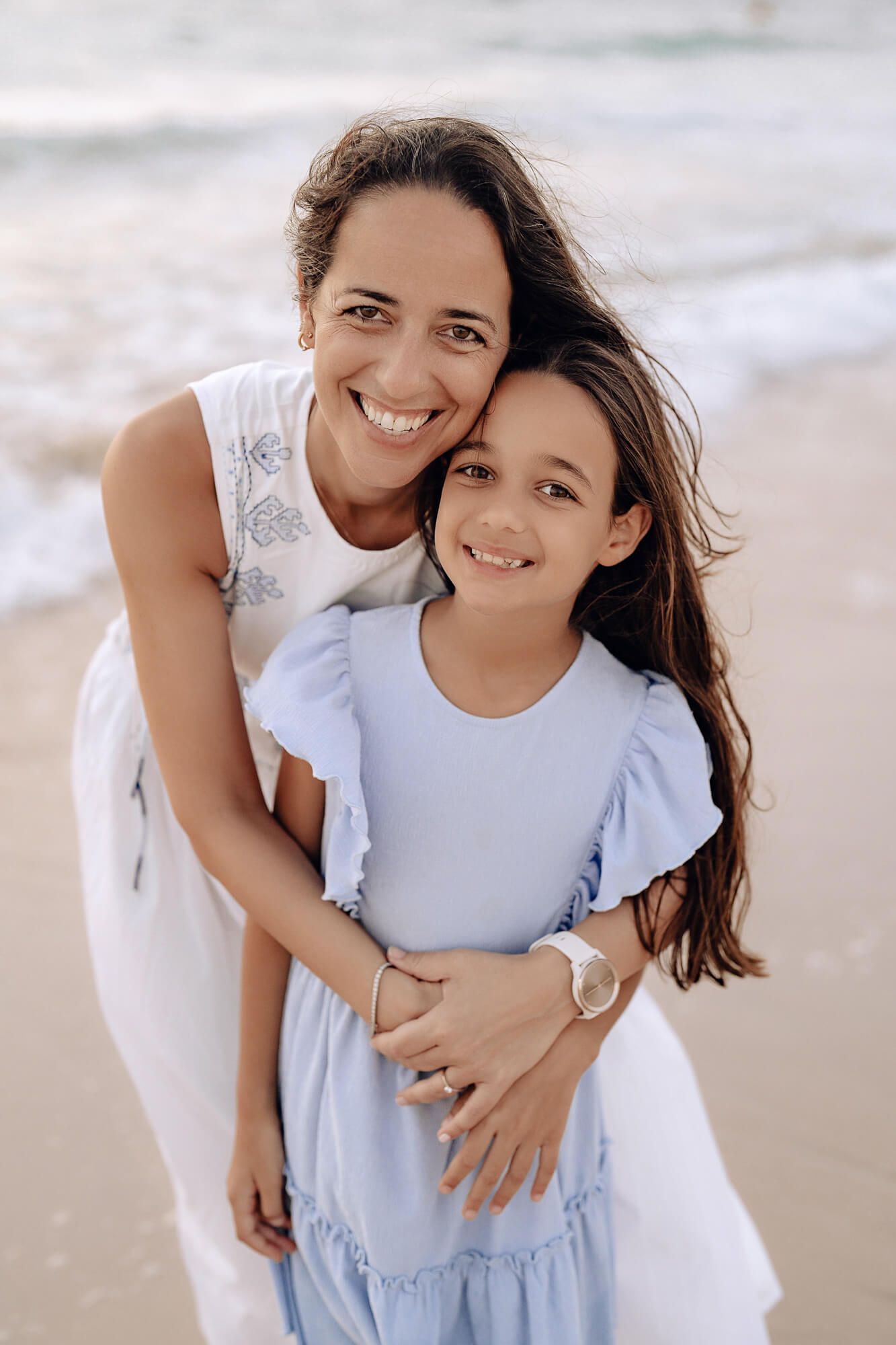 Candid beach portraits showing joyful embraces between two people in white and light blue outfits.