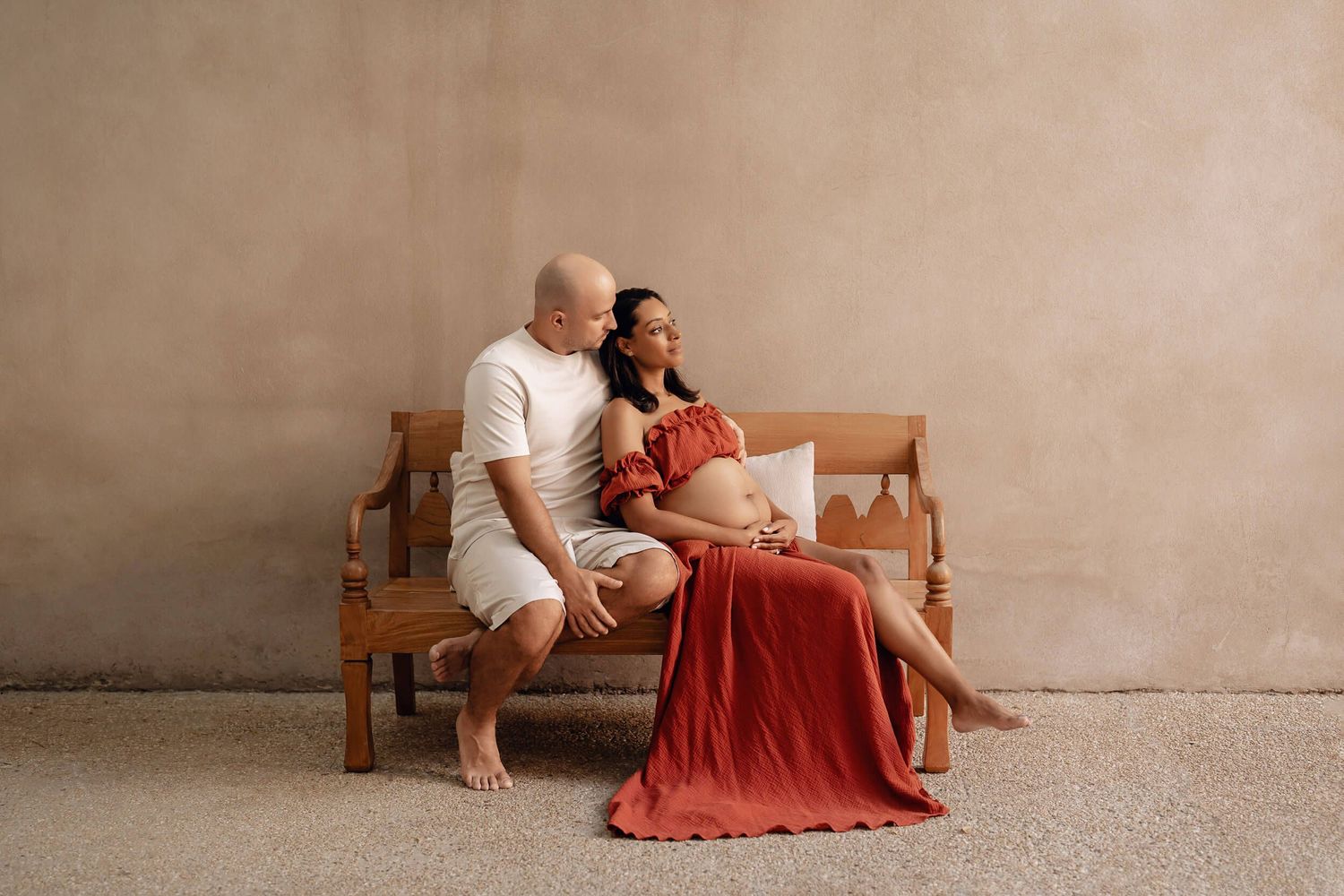 Intimate maternity session with couple sitting on wooden bench against neutral wall.