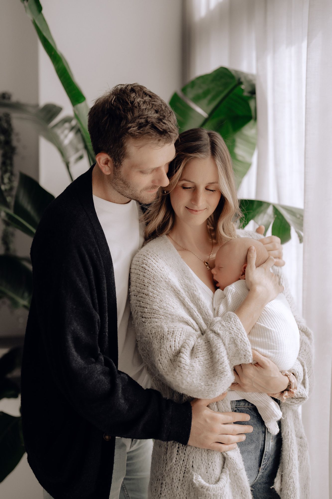 A couple shares an intimate moment standing together by a window with large tropical plants in the background.