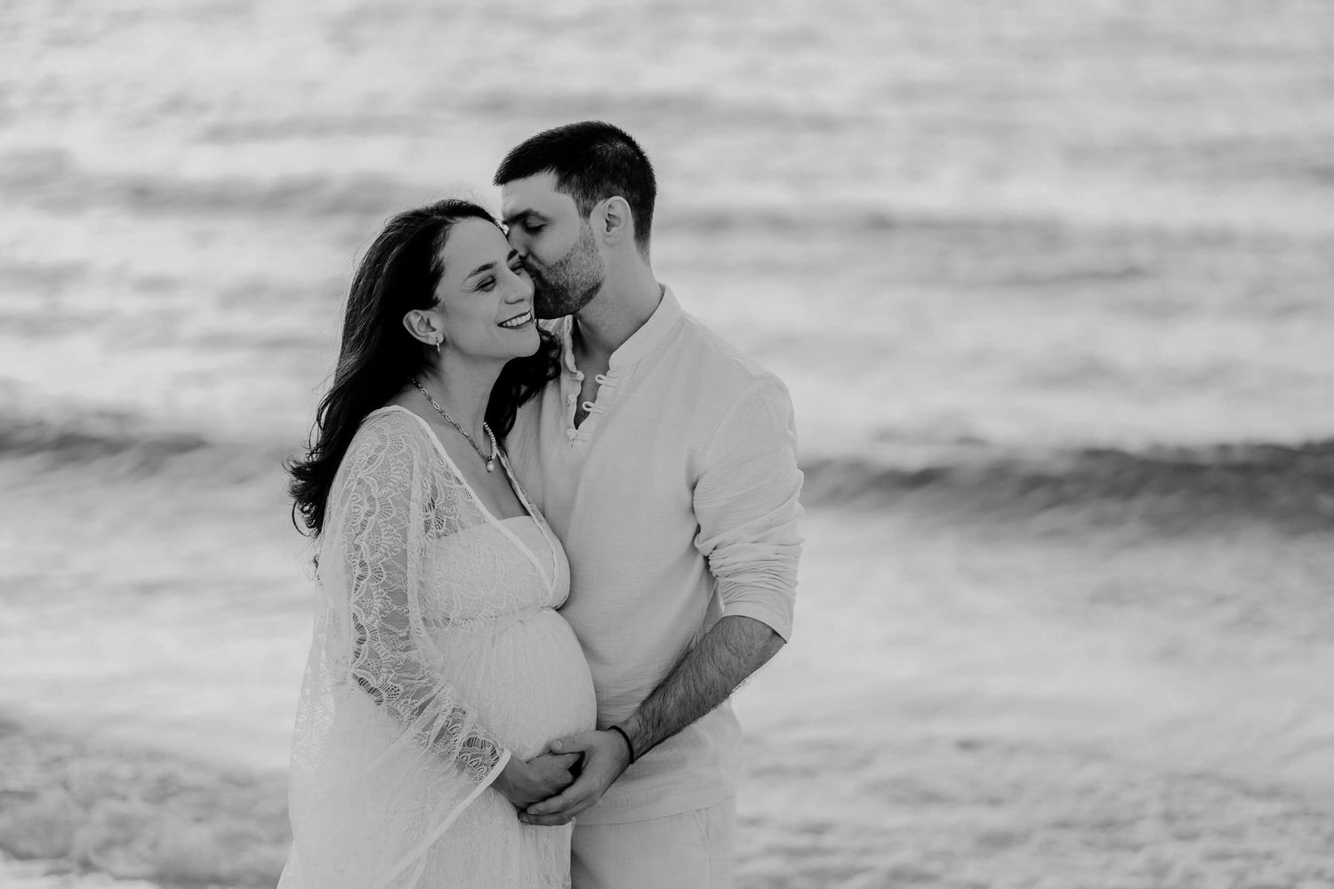 A romantic black and white photo of an expectant couple embracing by the ocean.
