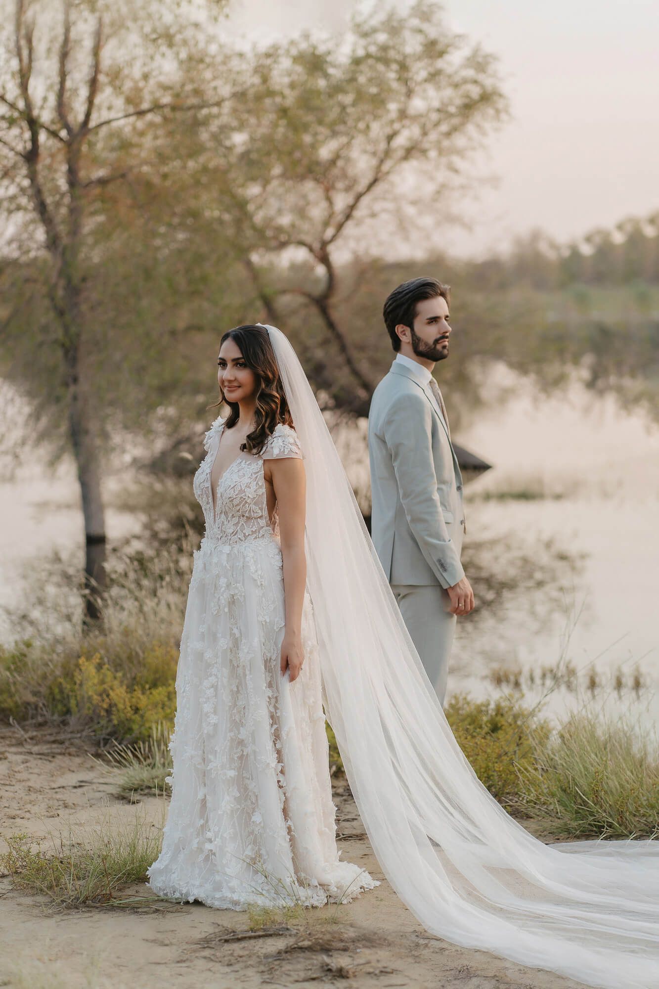 Bride in flowing lace dress and groom in gray suit stand back-to-back in a dreamy outdoor setting with long veil.