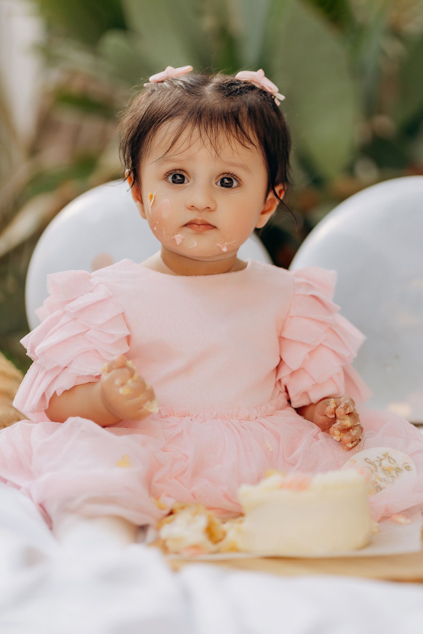 A baby wearing a ruffled pink dress sits on white cushions at a party.