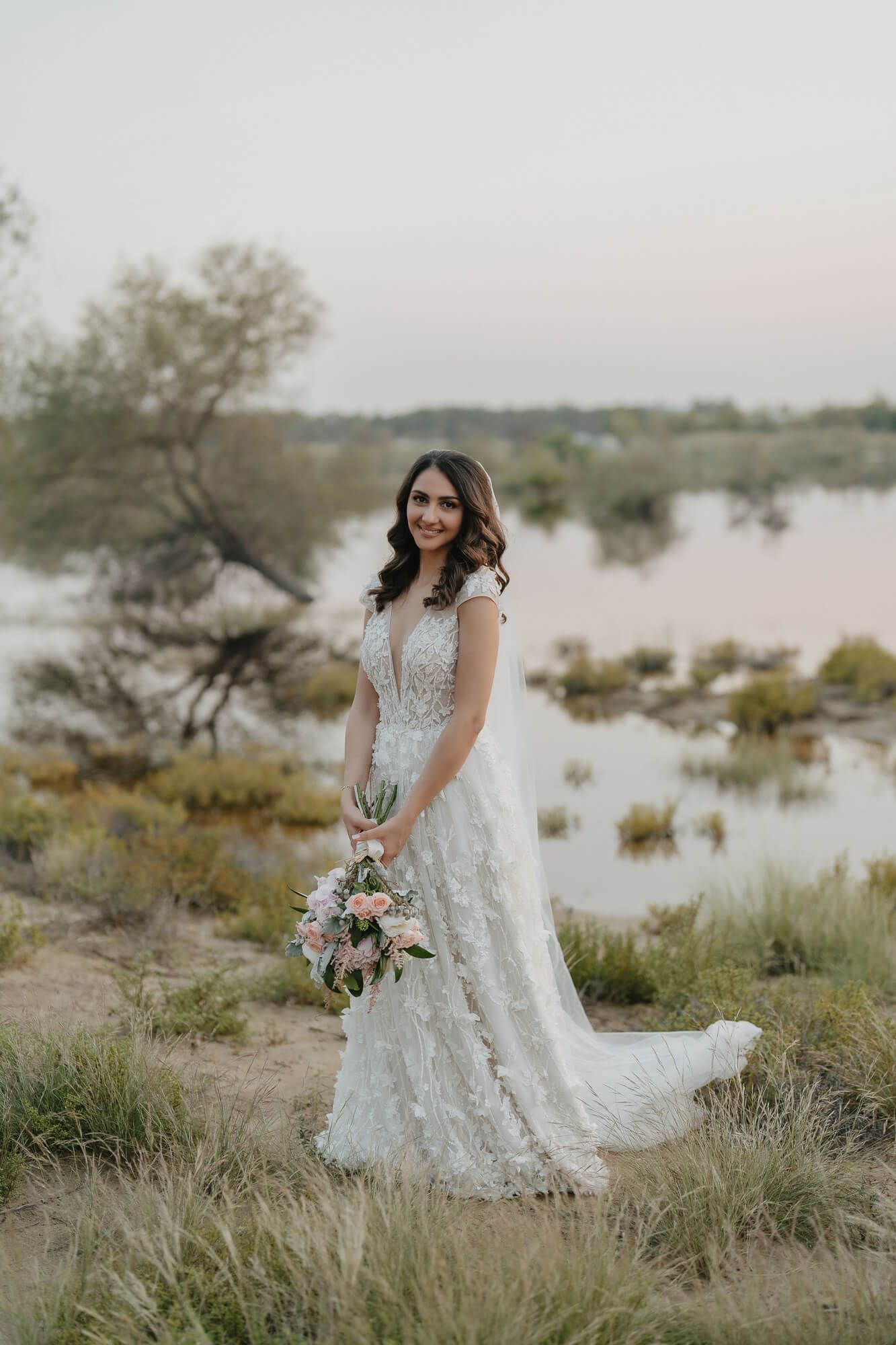 Bride in lace wedding dress holds bouquet while standing near lake at sunset with desert landscape in background.