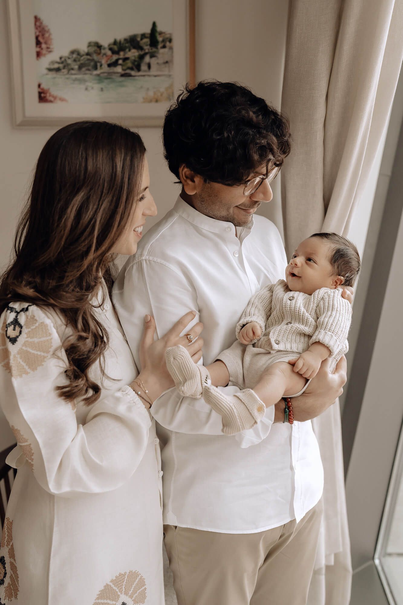Family in white outfits sharing a tender moment near a window with artwork visible.