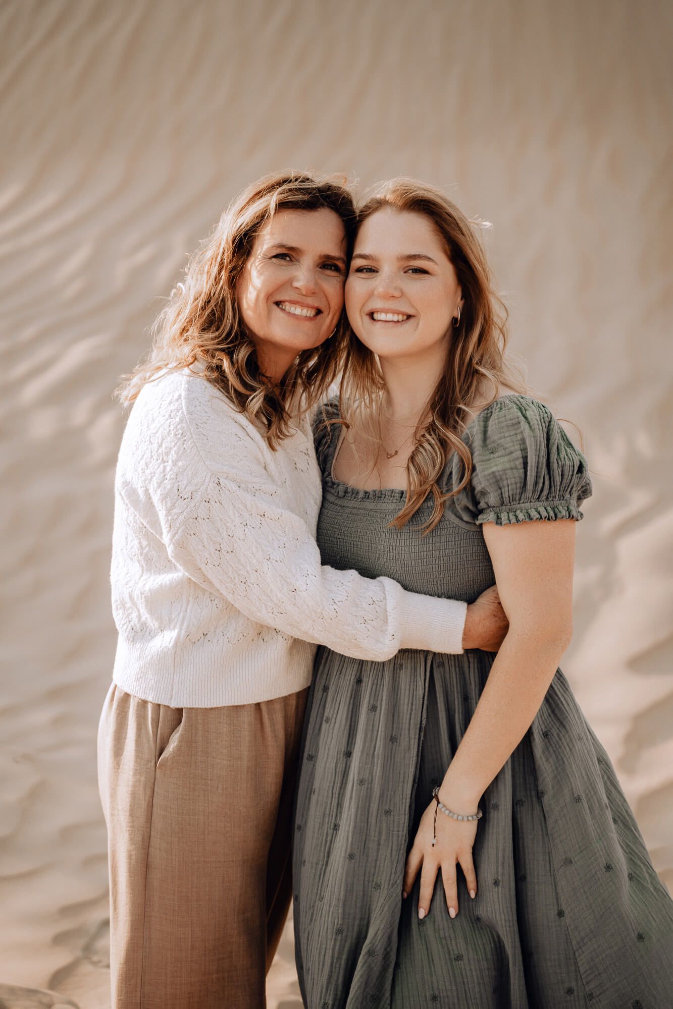 Two people embrace and smile warmly against sand dunes wearing casual earth-toned outfits.