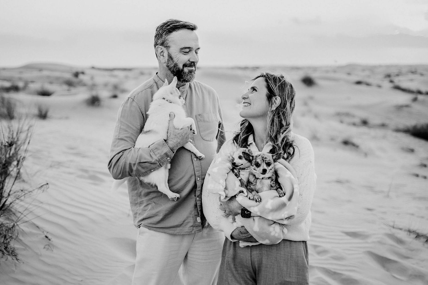 Joyful couple holding their small dog while sitting in desert sand dunes.