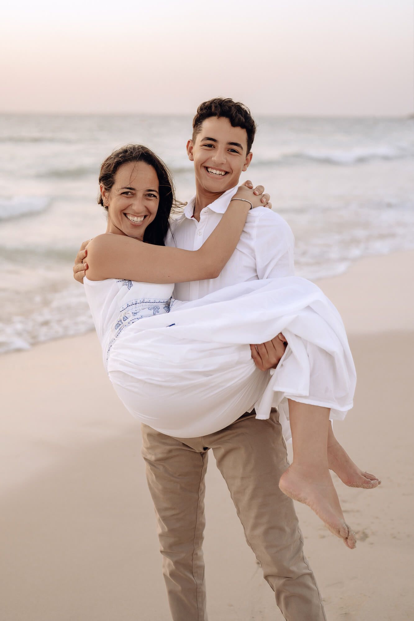 Two people in coordinated white and beige outfits share playful moments on a sandy beach.