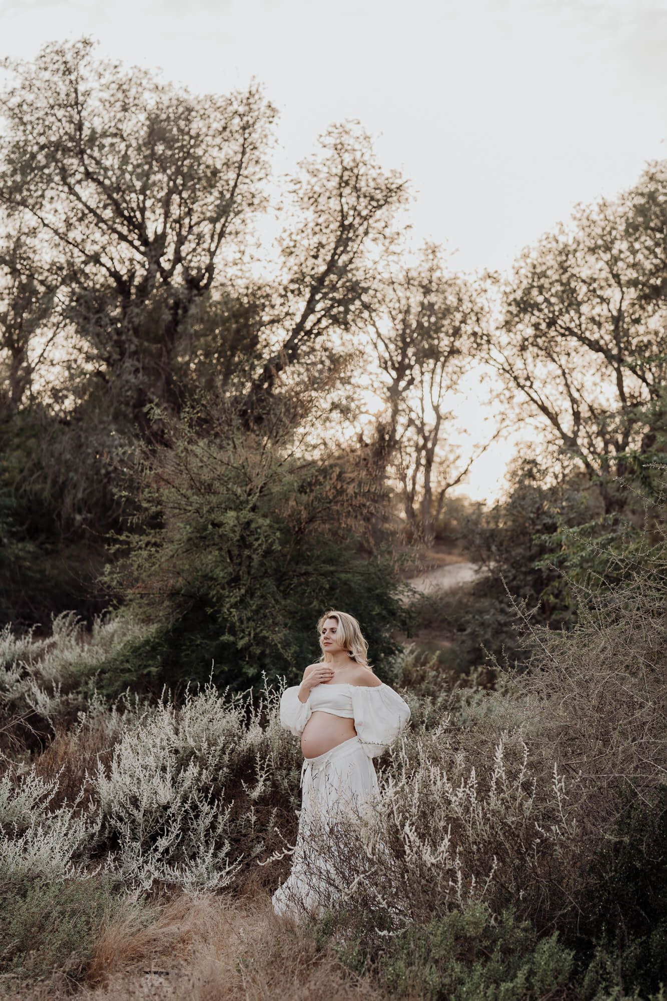 An expectant mother in a white dress poses in a natural outdoor setting at sunset.