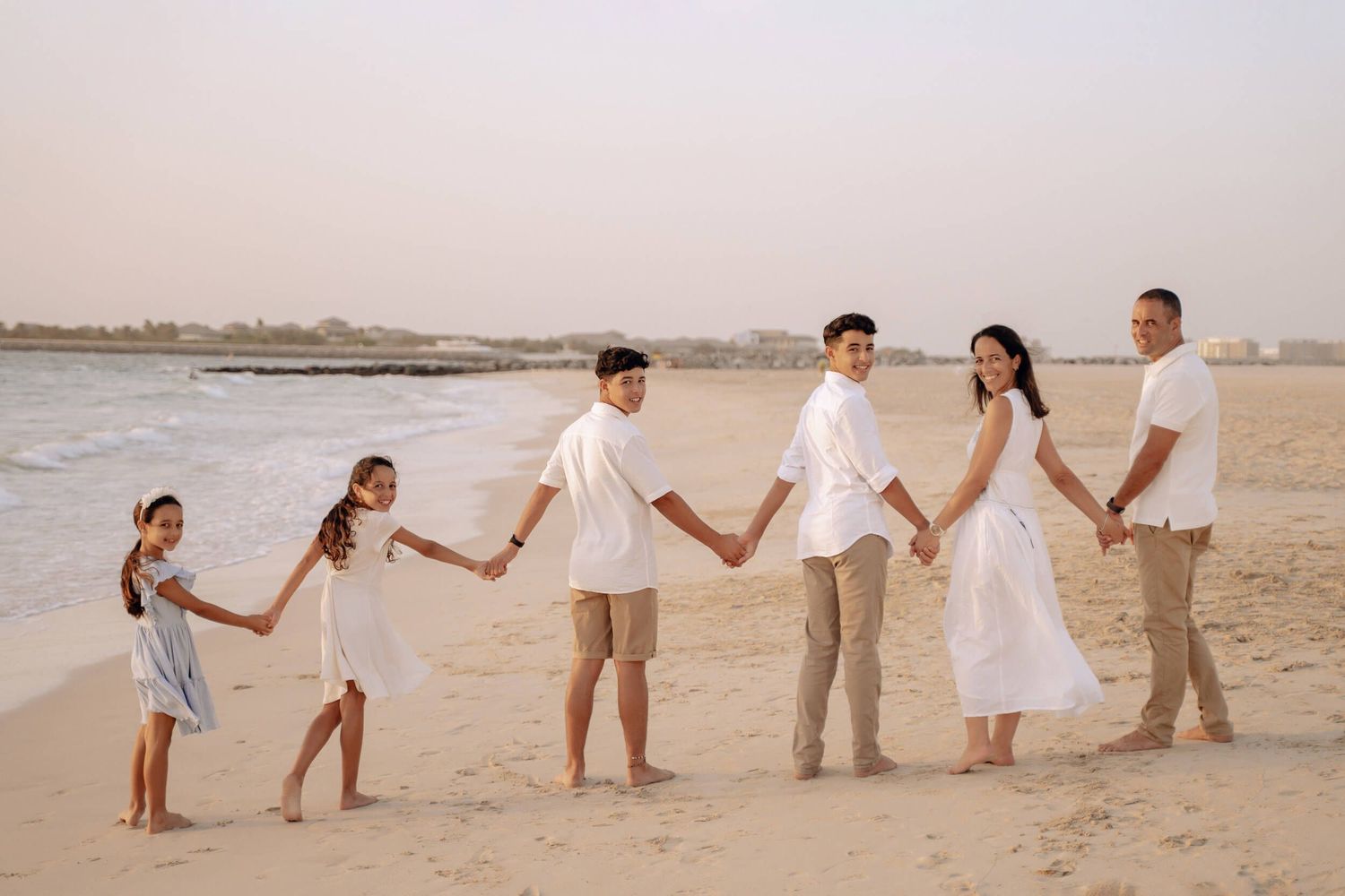 Family forms human chain on beach at sunset wearing coordinated white and beige outfits.