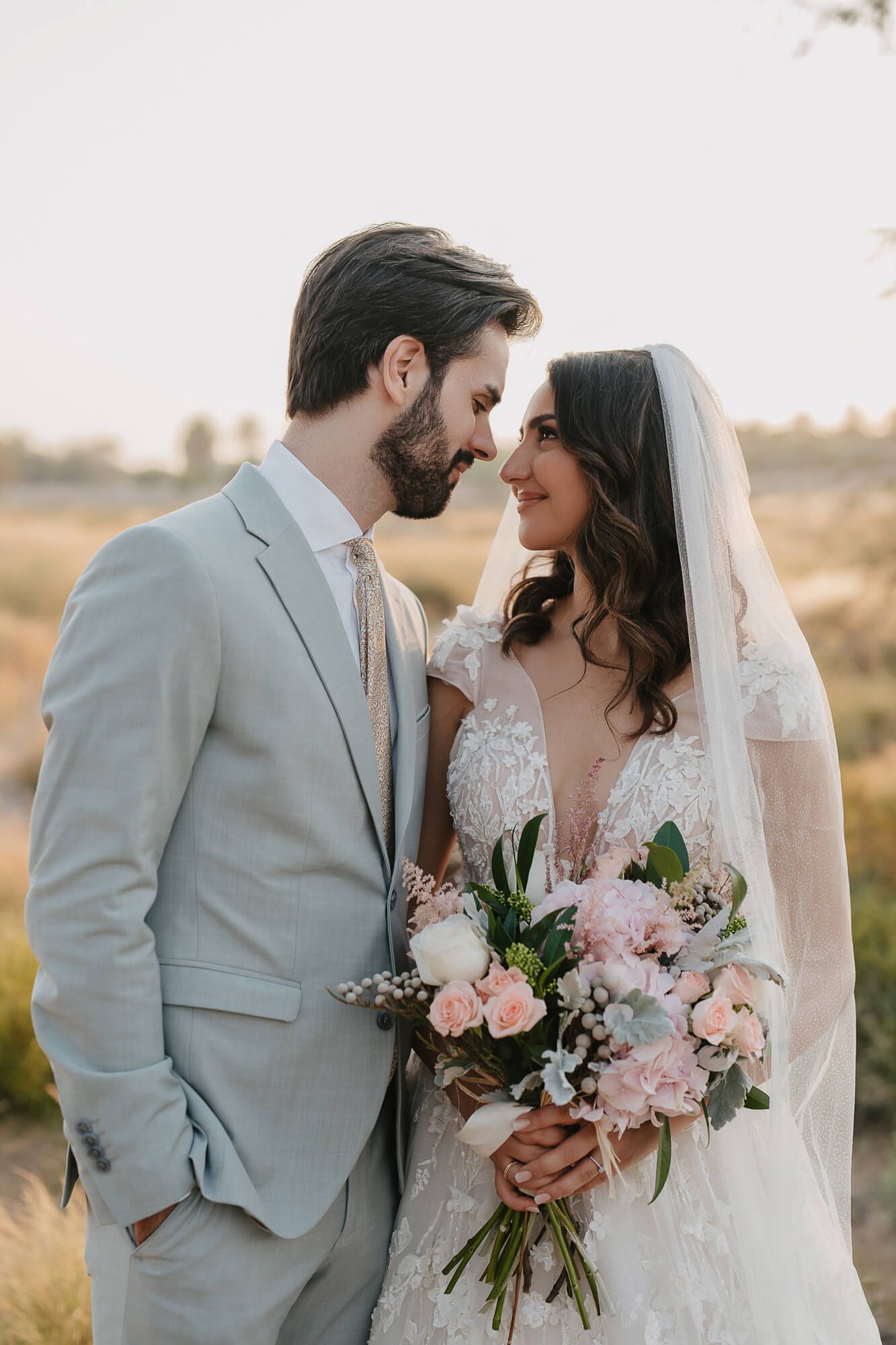 Romantic wedding portrait of couple in grey suit and white lace dress holding pink floral bouquet at sunset.