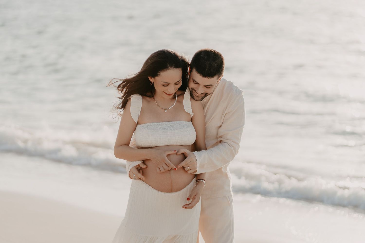 A couple on a sandy beach embraces tenderly during a maternity photoshoot, with waves in the background.