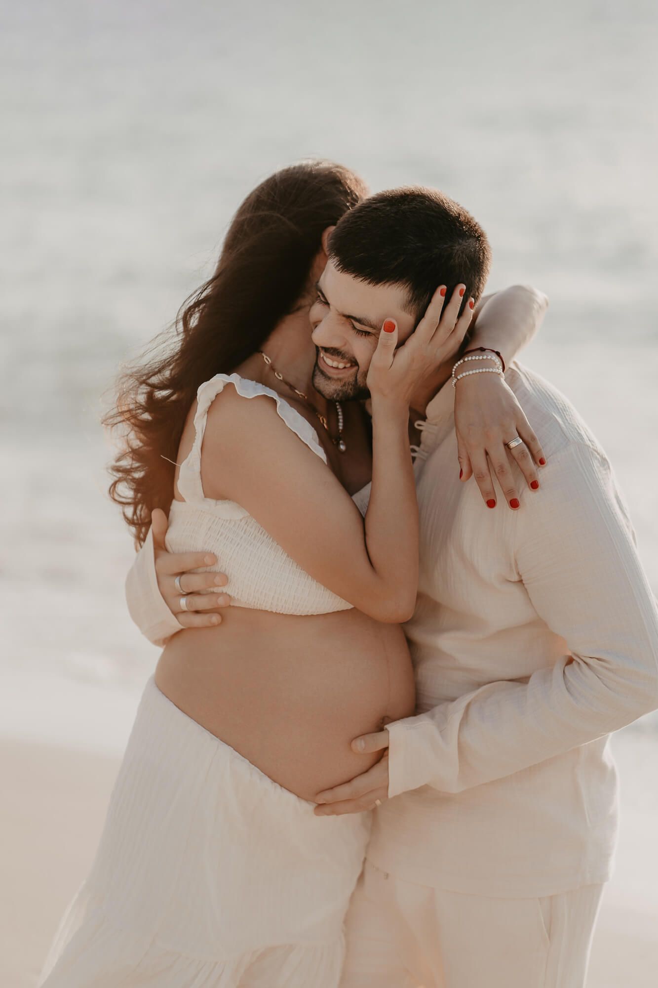 A tender maternity photo of a couple in white embracing on a beach during sunset, showing her pregnant belly.