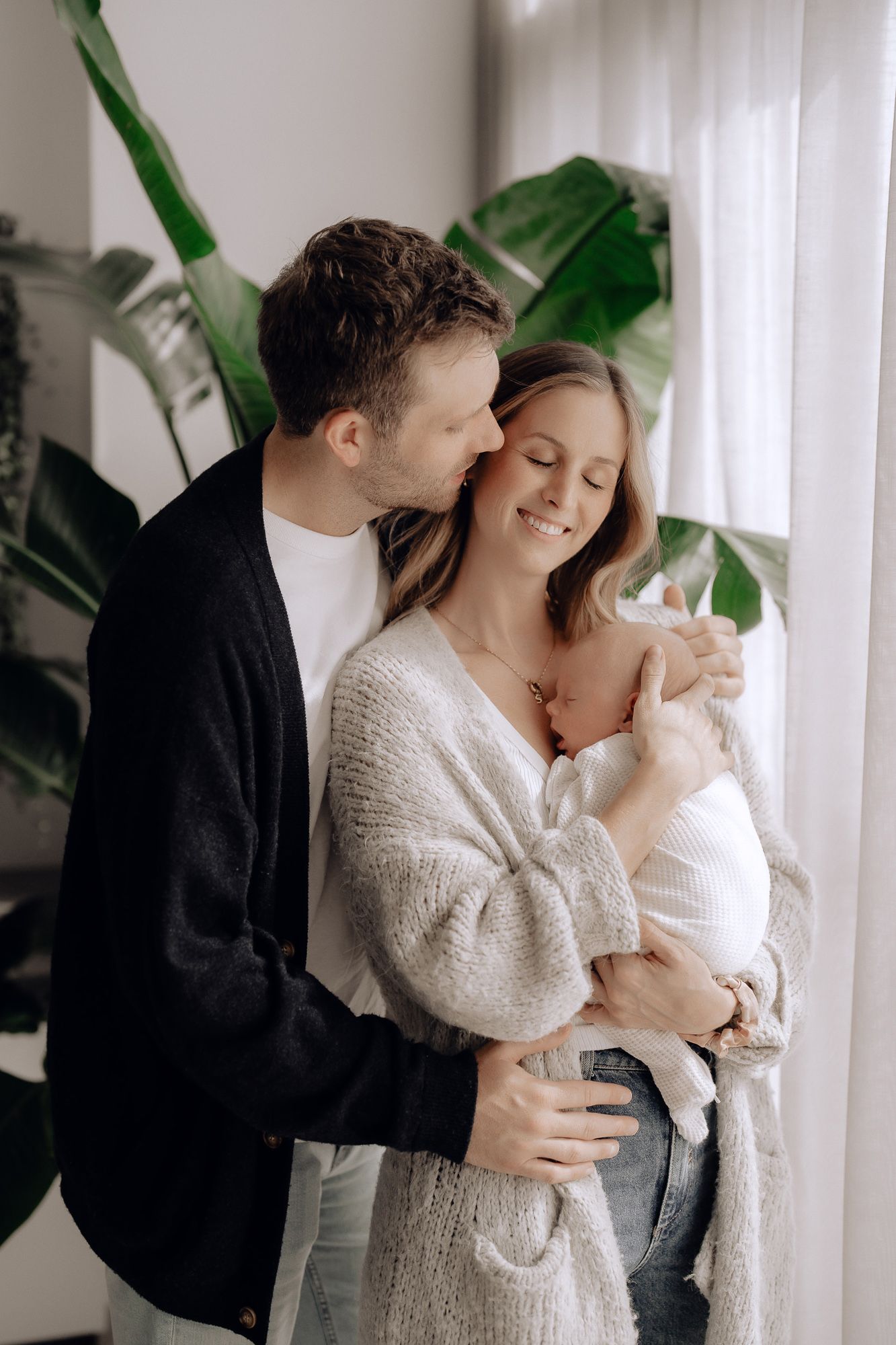 A couple embraces tenderly while holding their baby in a bright, minimalist room with potted plants.