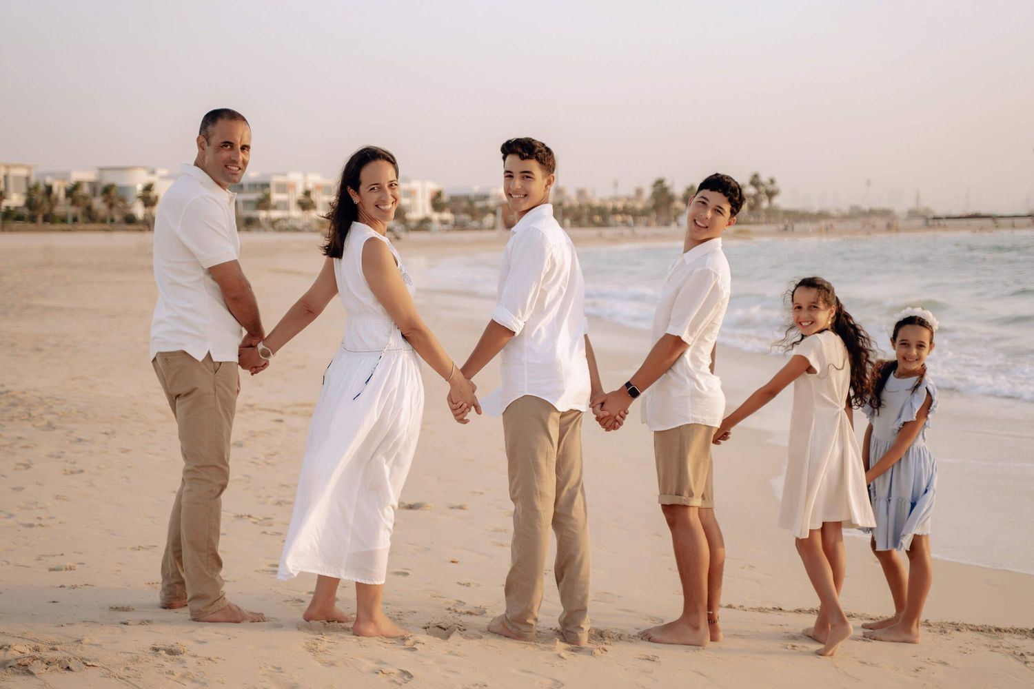 Family members lined up holding hands on beach in coordinated white outfits during sunset.