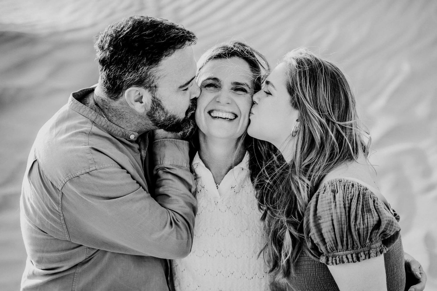 Black and white candid photo of three people sharing joyful kisses and laughter outdoors.