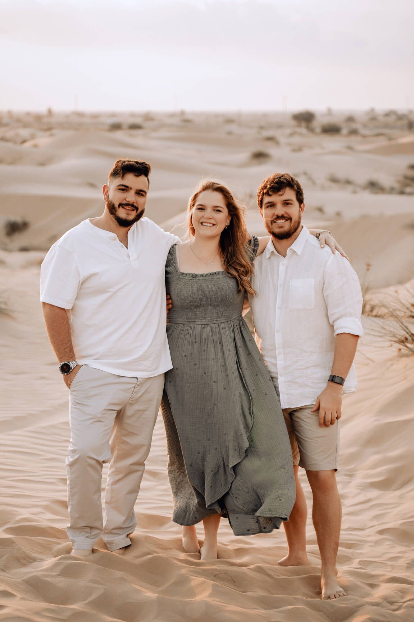 Three people in coordinated outfits pose together on desert sand dunes during sunset.