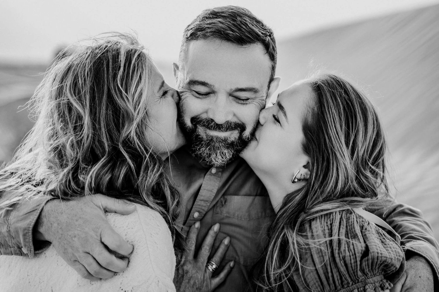 Black and white portrait capturing tender moment of three people embracing outdoors.