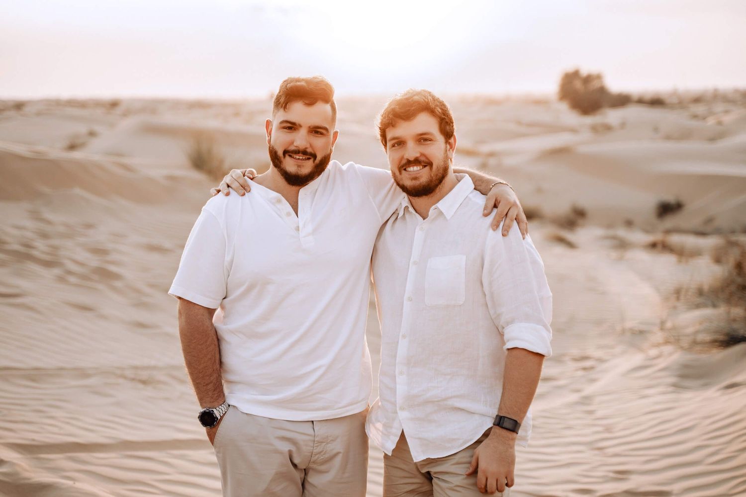 Two people smiling while wearing matching white shirts against desert sand dunes at sunset.