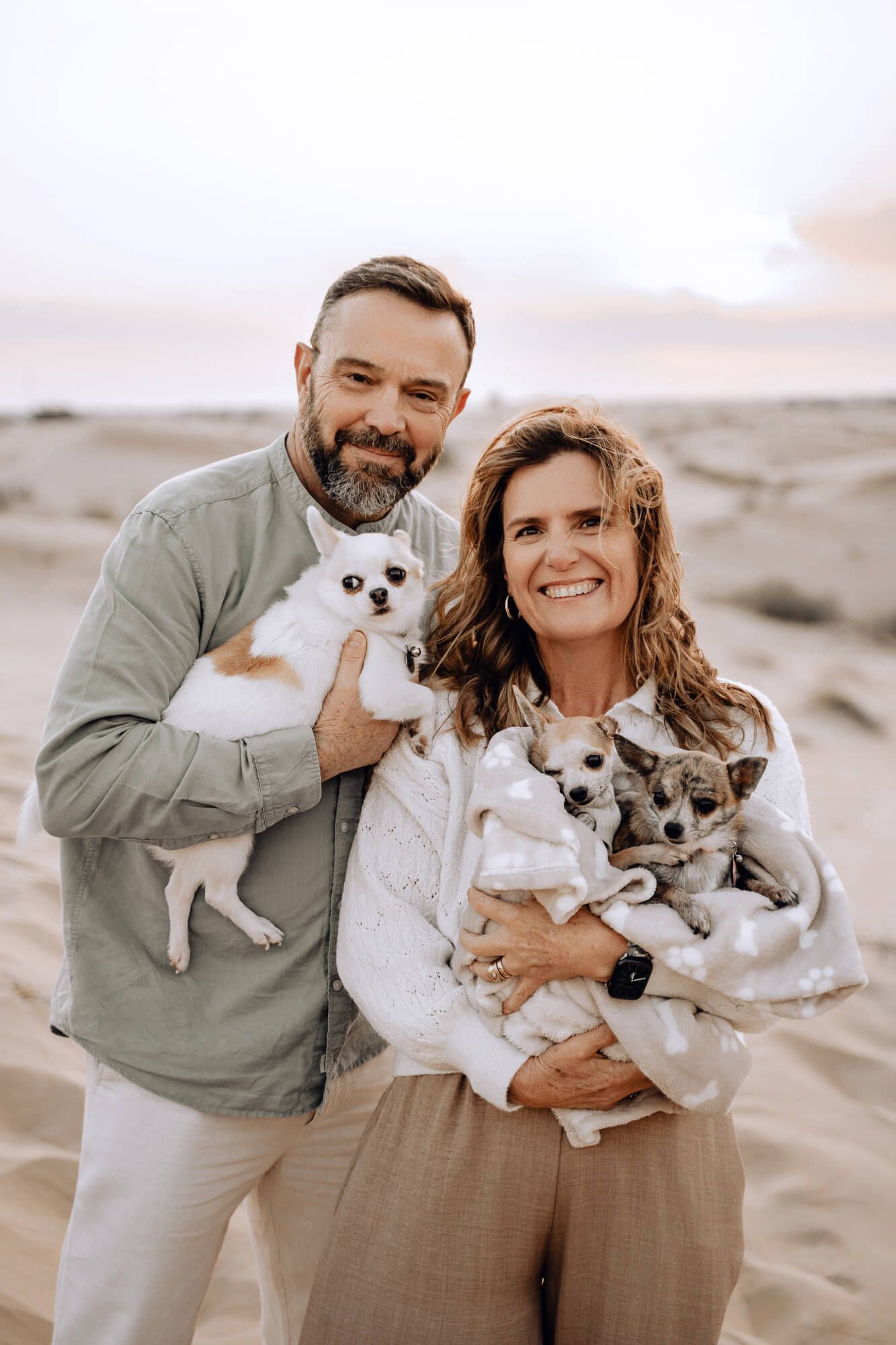 Couple holding small white dogs while posing on desert sand dunes at sunset.