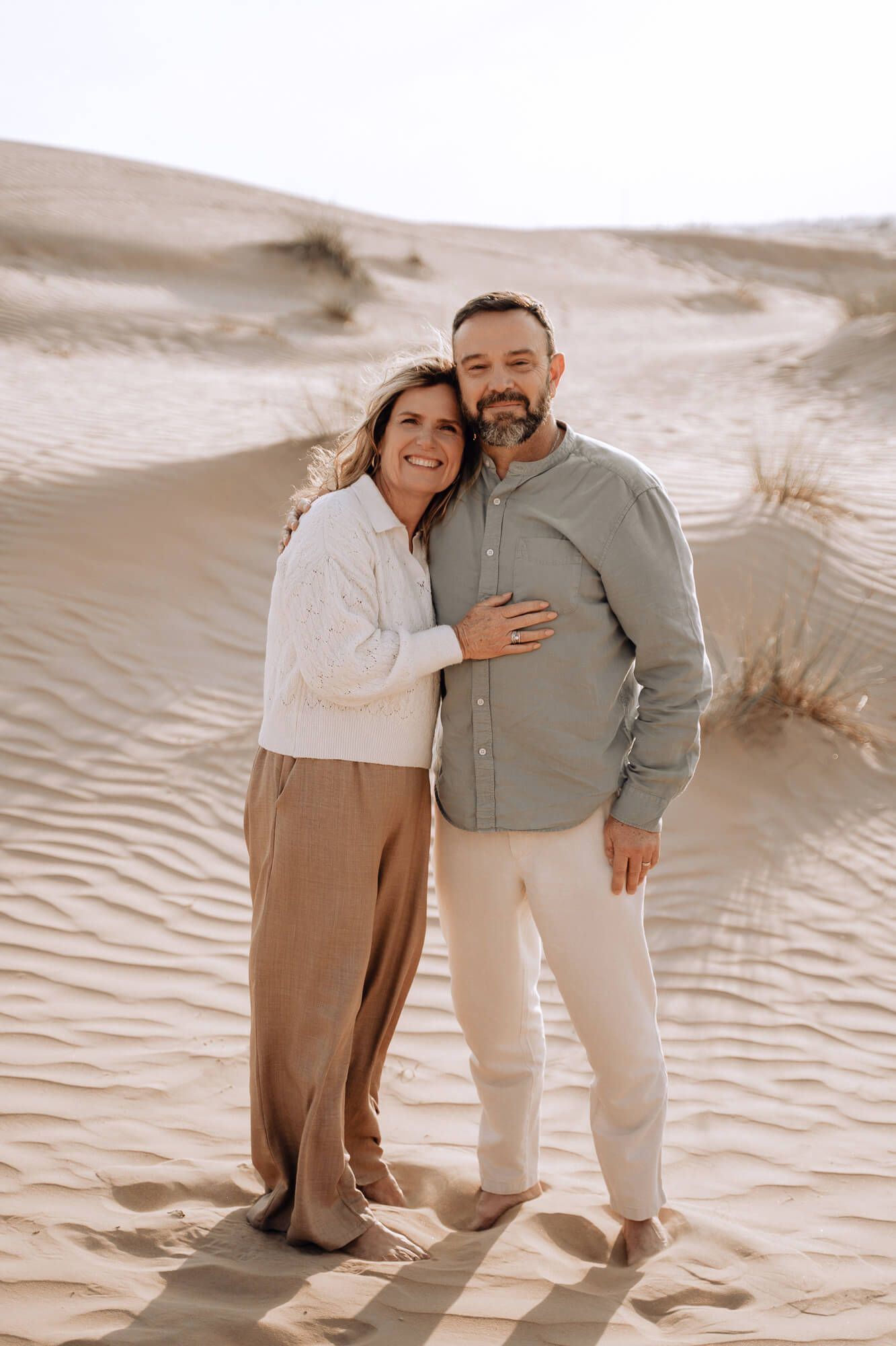 Couple in neutral tones sharing intimate moments while walking through sand dunes during a photoshoot.
