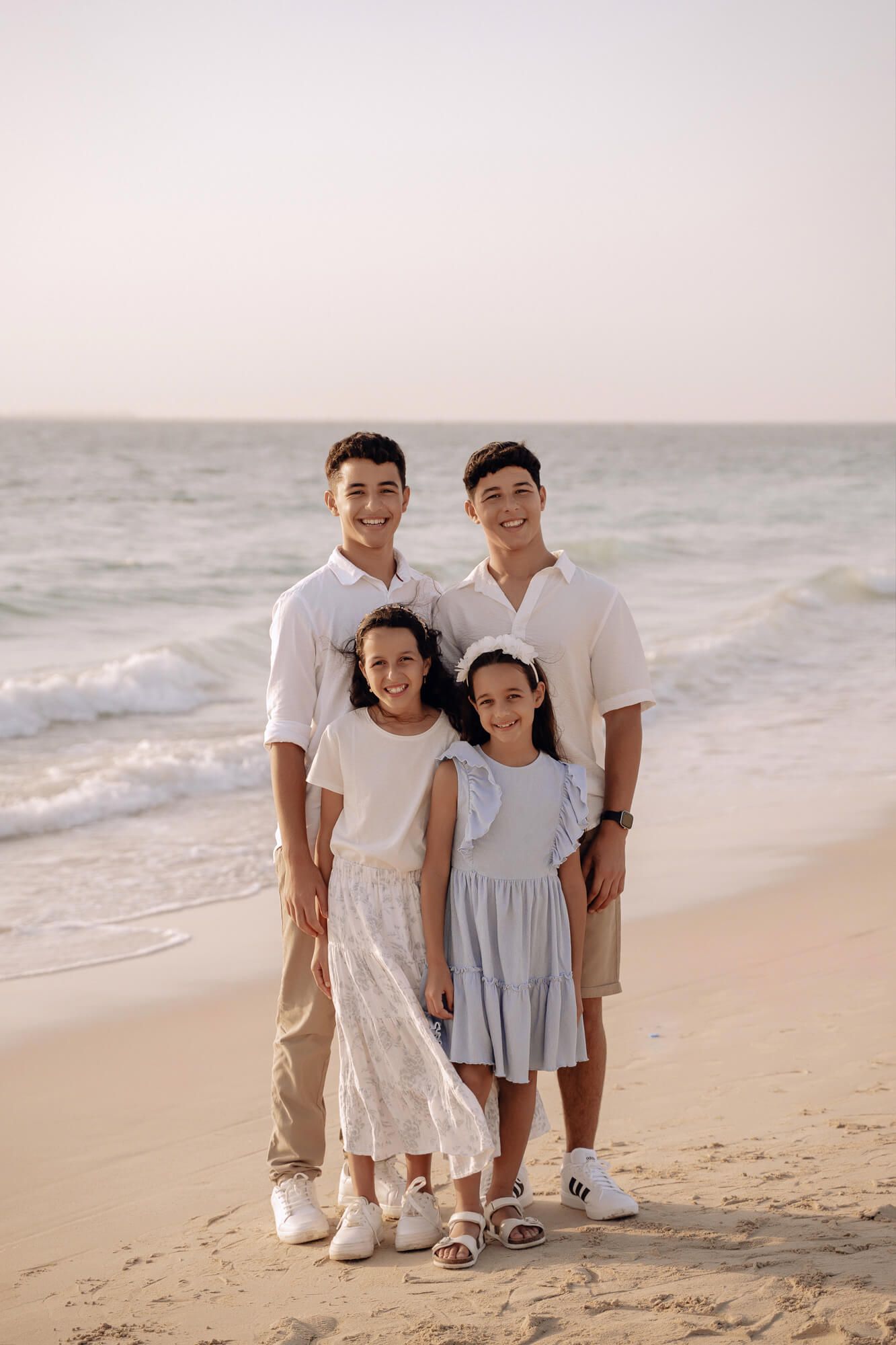 Family of four dressed in coordinating white outfits poses together on a beach at sunset.