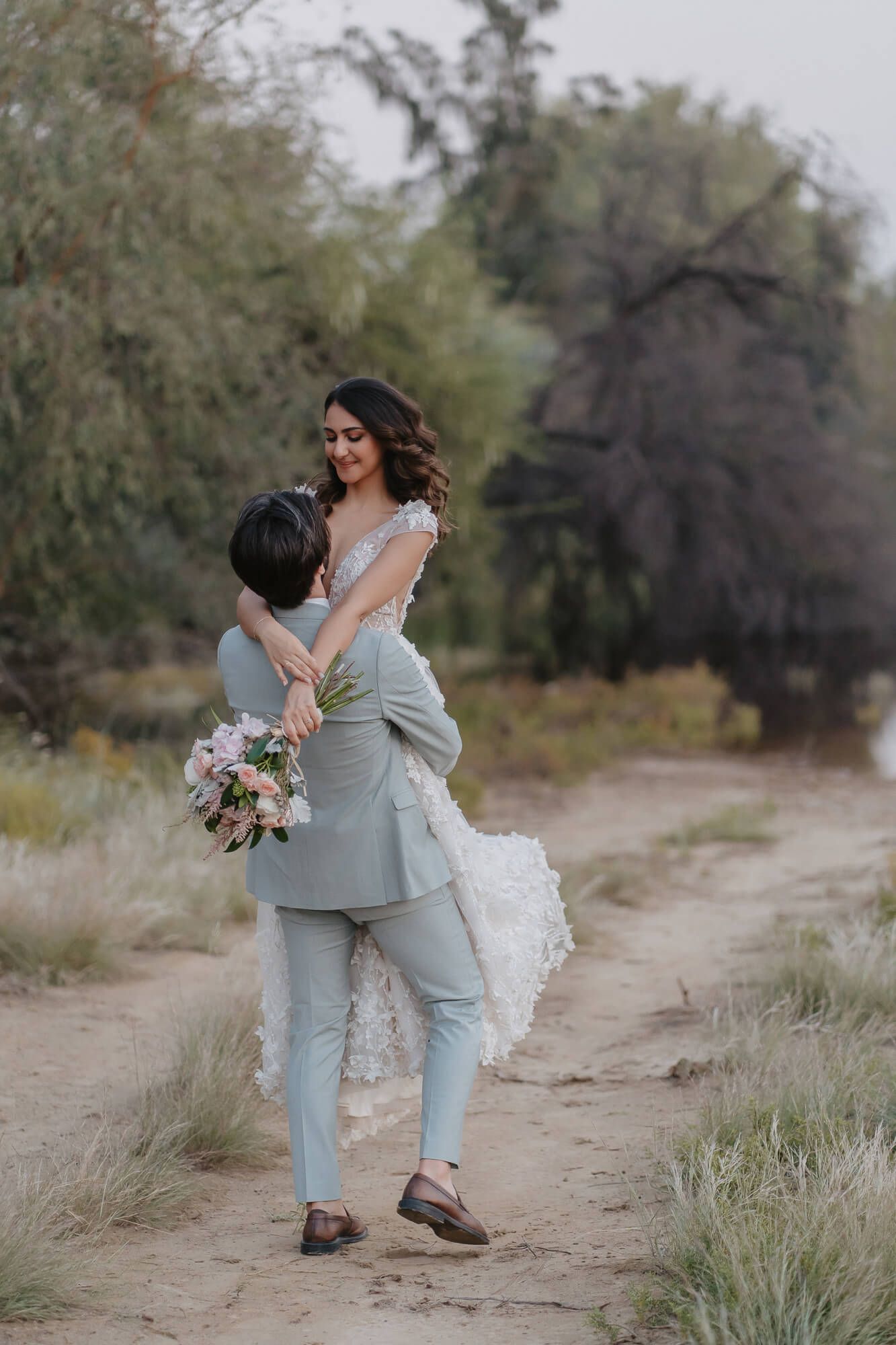 A romantic wedding couple embraces on a dirt path surrounded by natural greenery while holding a floral bouquet.