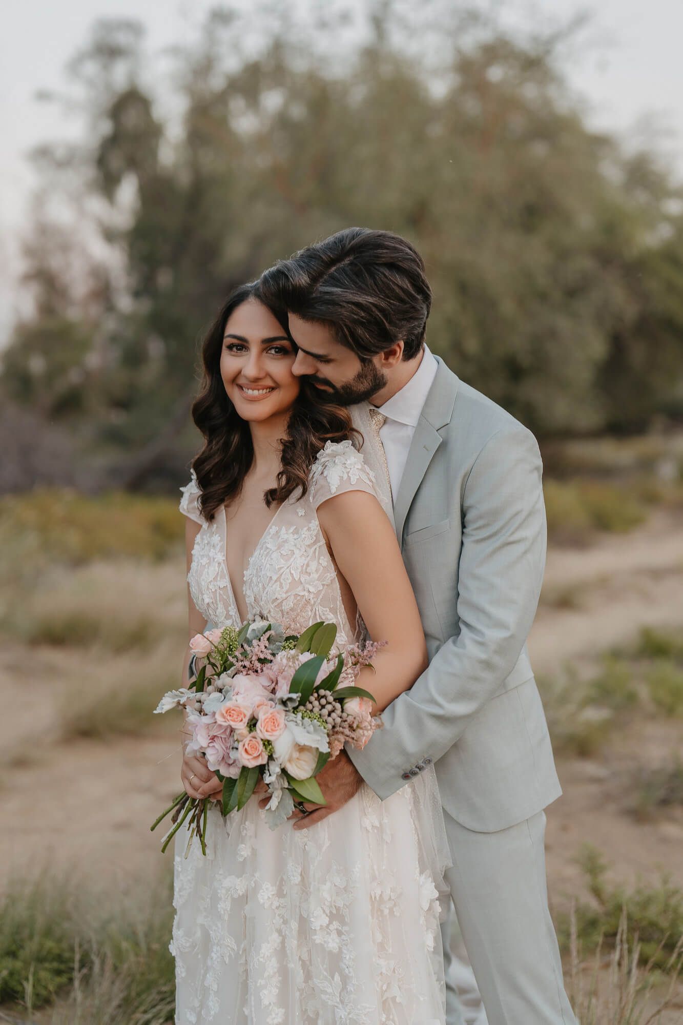 A romantic wedding couple embraces in a desert landscape wearing light gray and white attire with a floral bouquet.